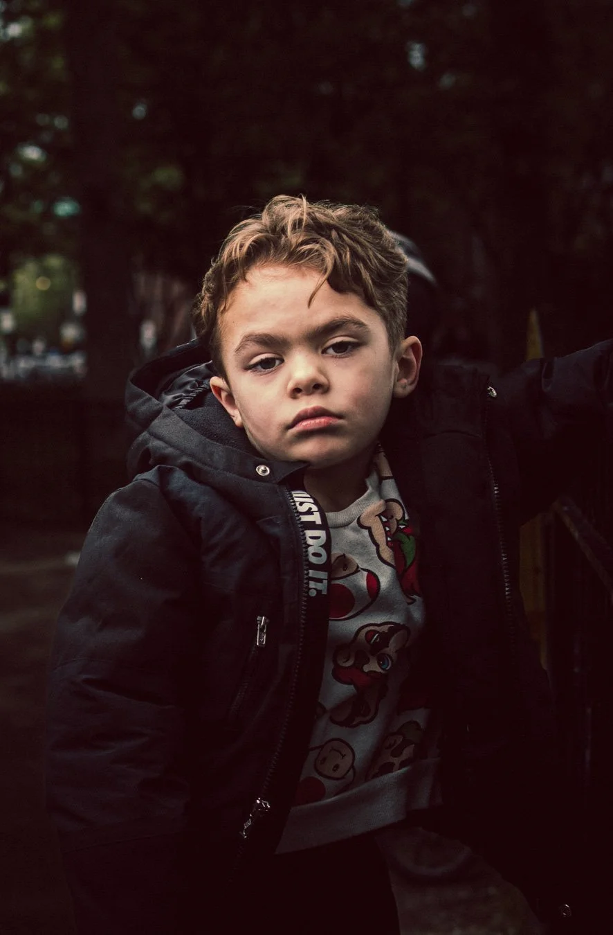 A young boy with brown hair and a serious expression, wearing a black jacket and a Mario-themed shirt, sitting outdoors with a dark background of trees.