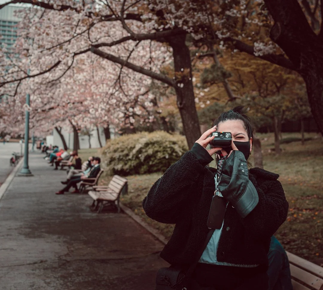 A woman taking a photograph with a camera in a park with pink blossom trees and benches, some people sitting and walking in the background.