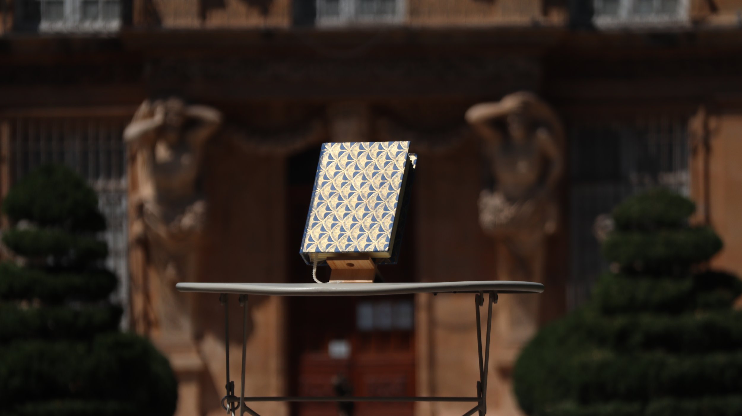 Une œuvre d'art japonaise sur un support en bois, avec un motif géométrique bleu et doré, exposée à l'extérieur avec des sculptures en bois et des arbres taillés en forme.