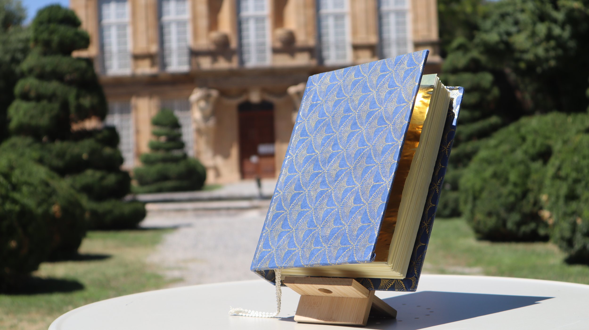 Livre avec une couverture bleue ornée d'un motif doré, placé en équilibre sur un support en bois, avec un jardin et un grand bâtiment historique en arrière-plan.