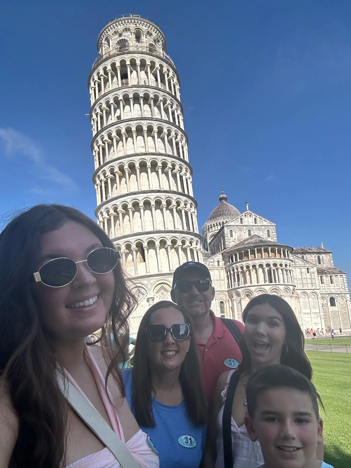 Group of five people posing for a selfie in front of the Leaning Tower of Pisa on a sunny day.
