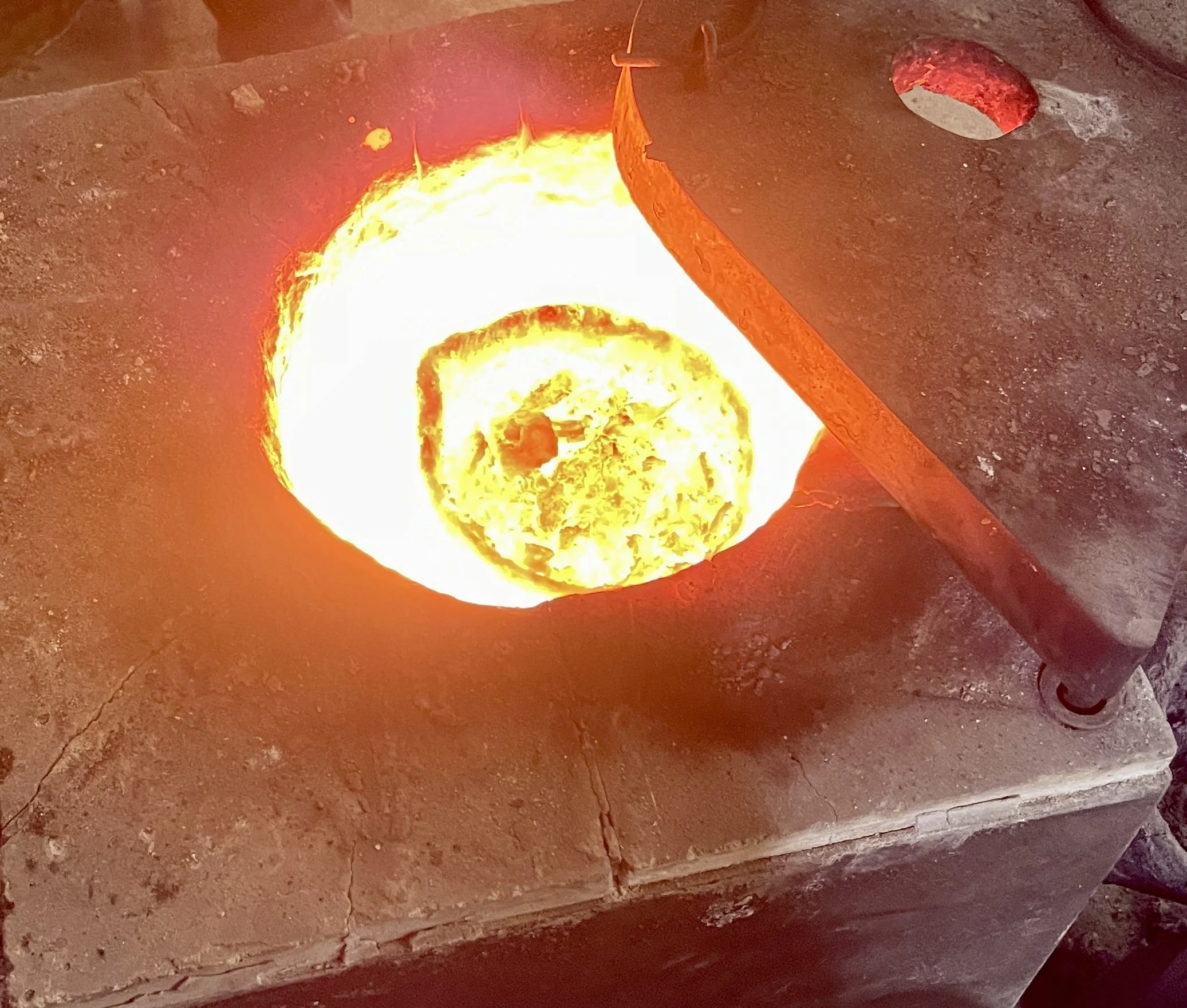 A glowing ball of molten metal in a forge or foundry, with a bright yellow and orange hue, partially enclosed by a metal structure.