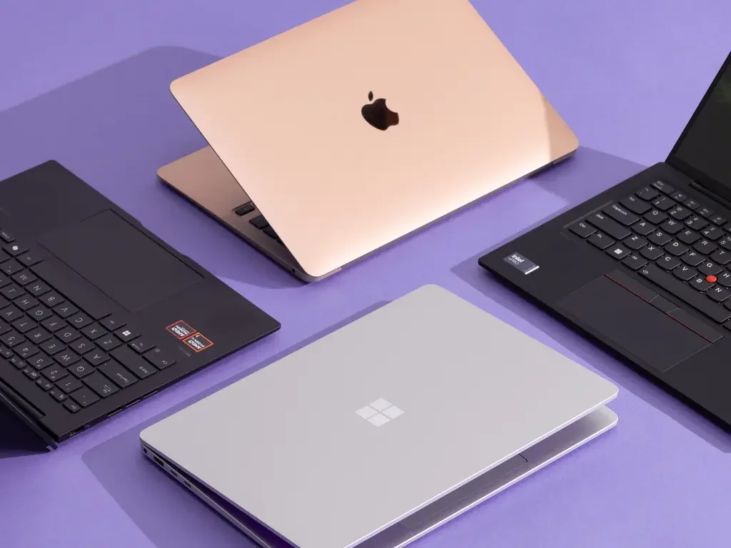 Four laptops on a purple surface, including a pink MacBook, a black Windows laptop, a gray Surface laptop, and a black ThinkPad.