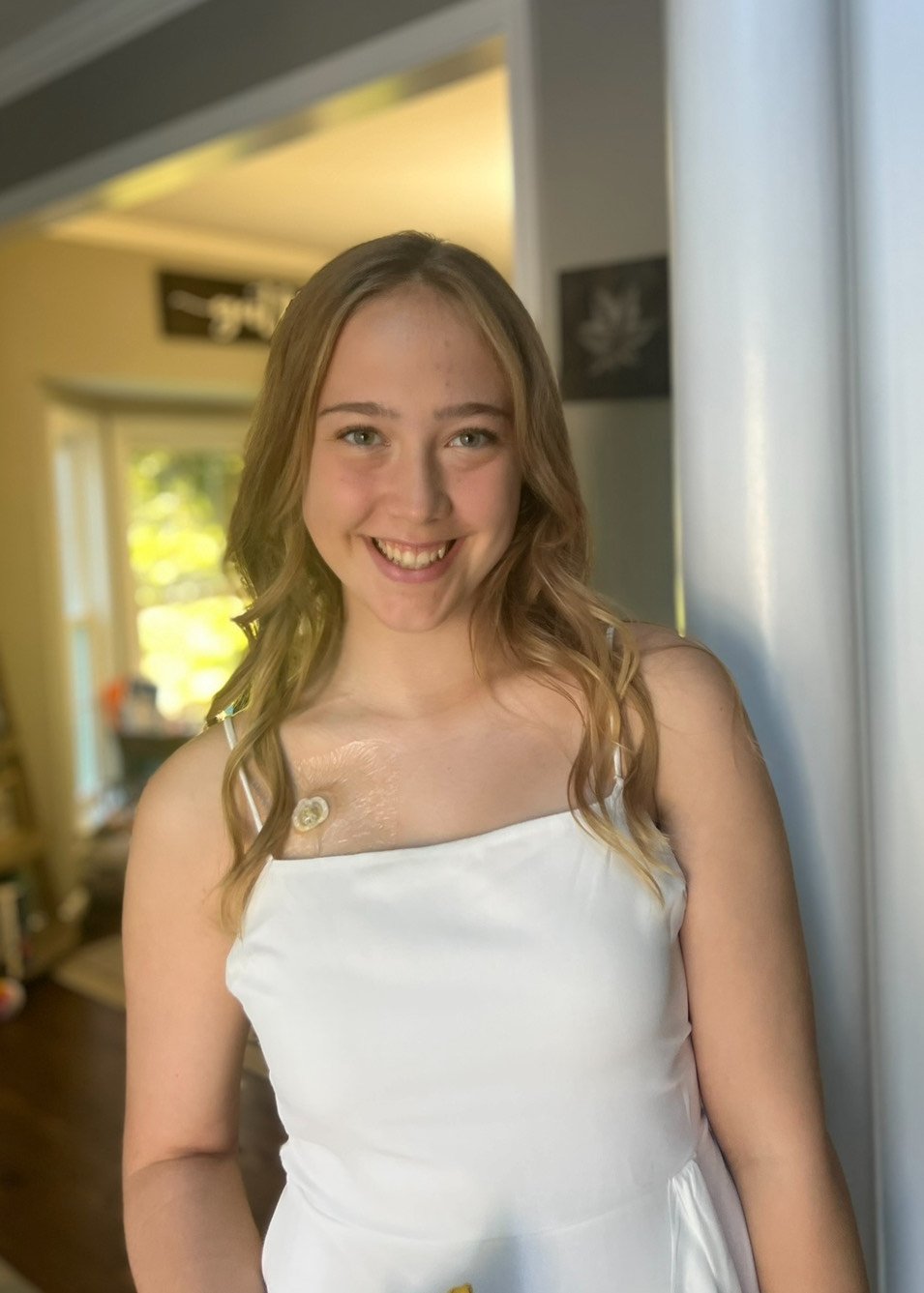 Young girl with green eyes and long, pink-tinted hair smiling while sitting in a car. She is wearing a cream-colored top, a colorful beaded necklace, and a name tag that reads 'Alexa.'