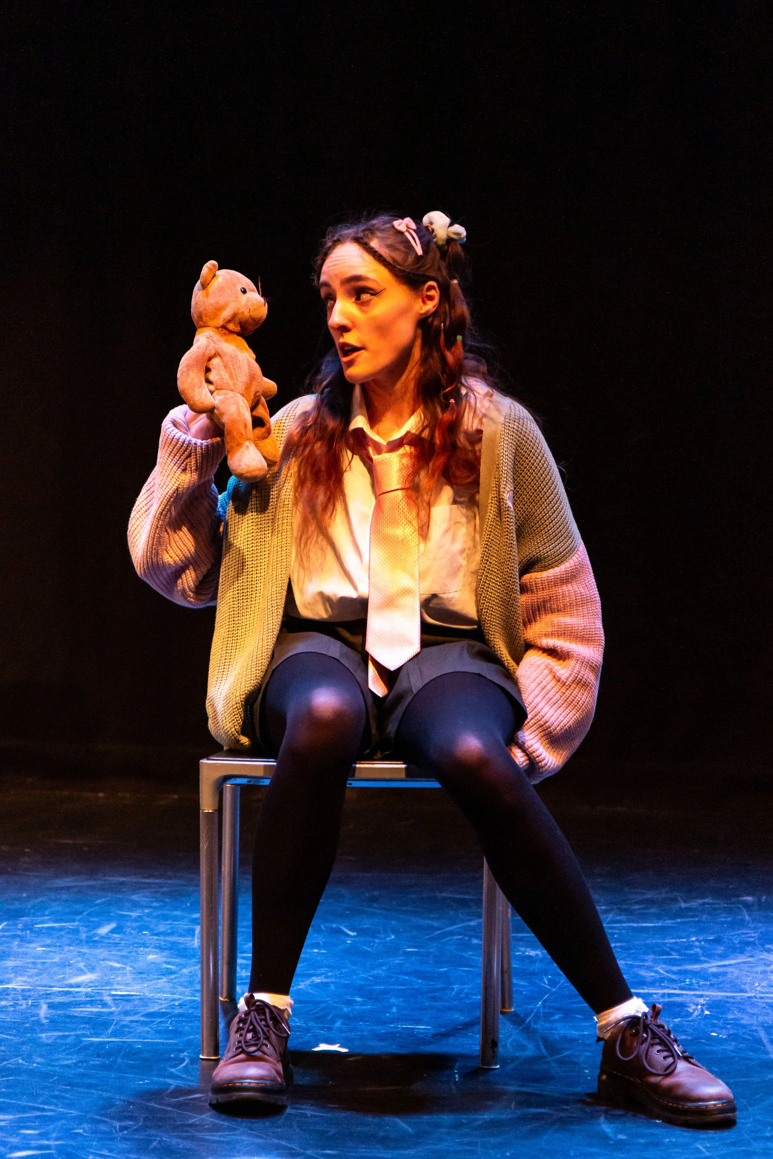 A woman with long wavy hair, wearing a beige blazer, white shirt, black tights, and brown boots, sits on a chair on stage. She holds a teddy bear near her face, looking at it with a thoughtful expression. The stage is dark with a blue-lit floor.