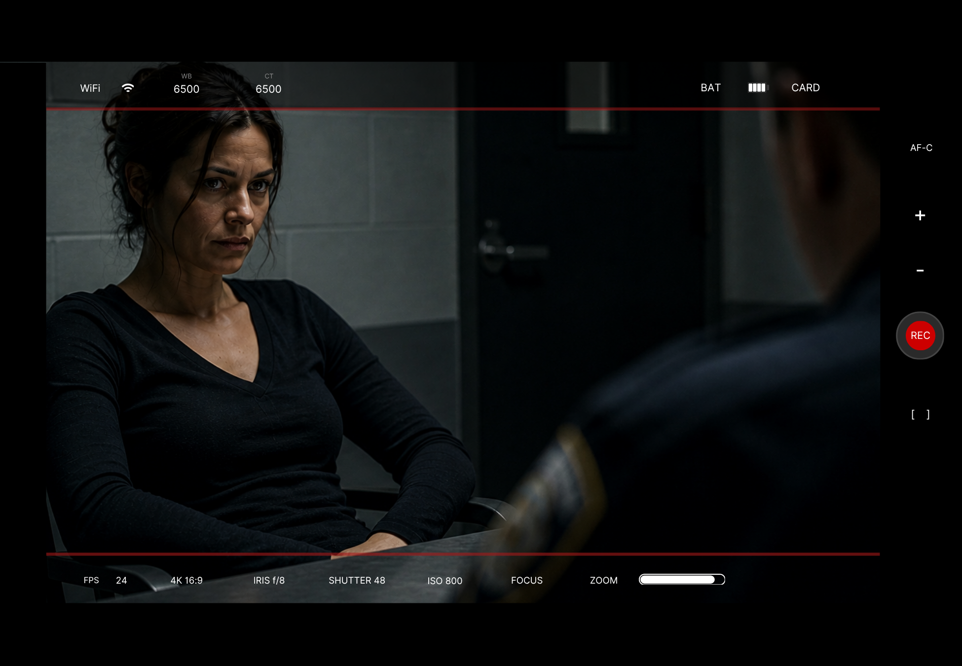 Woman with dark hair and tanned skin wearing a black long-sleeve shirt, sitting in a gray room with a concerned expression, facing another person whose face is partially visible.