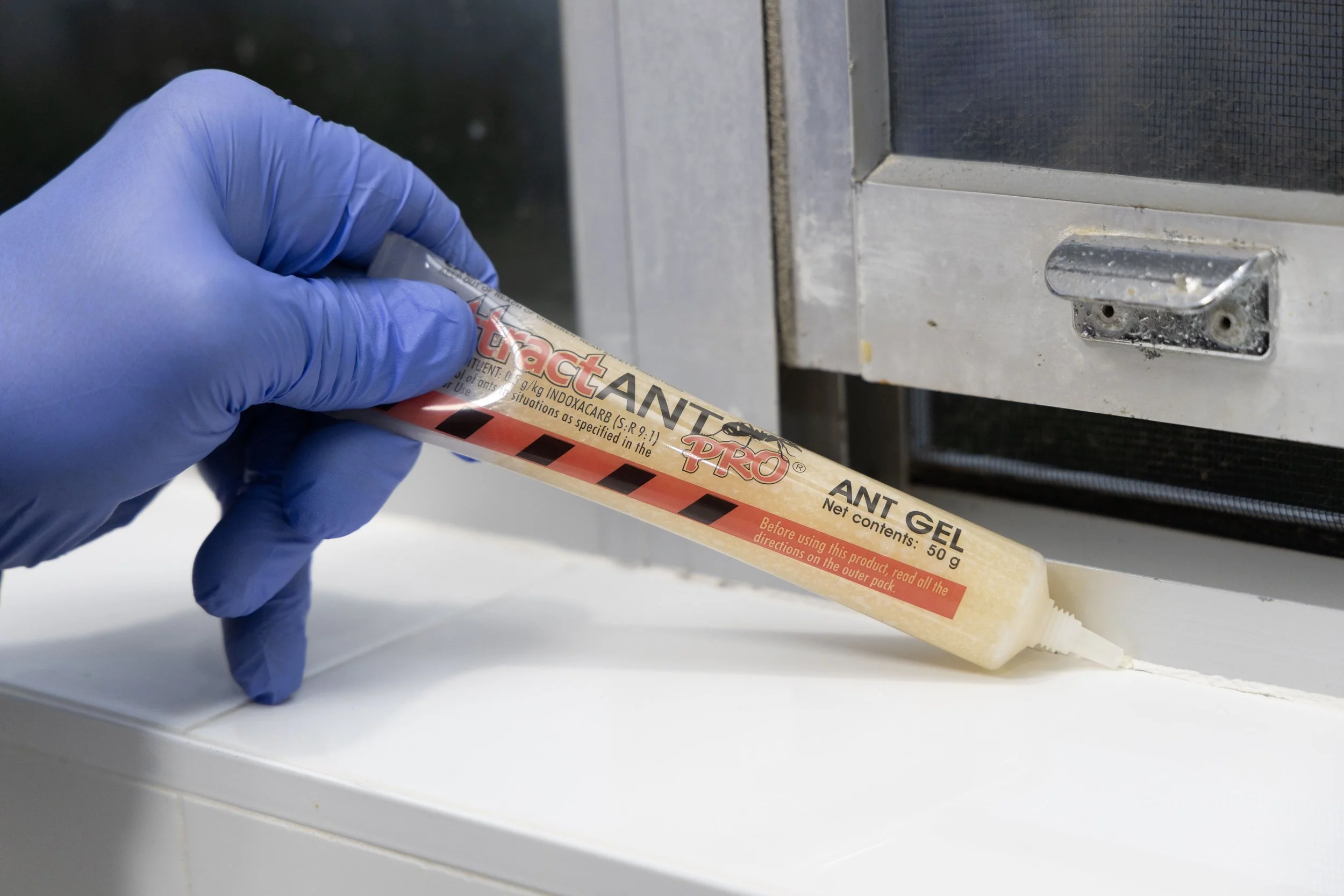 A person wearing a blue glove holding a tube of ANT GEL insect bait, with the nozzle pressed against the edge of a white surface, near a metal appliance or cabinet.