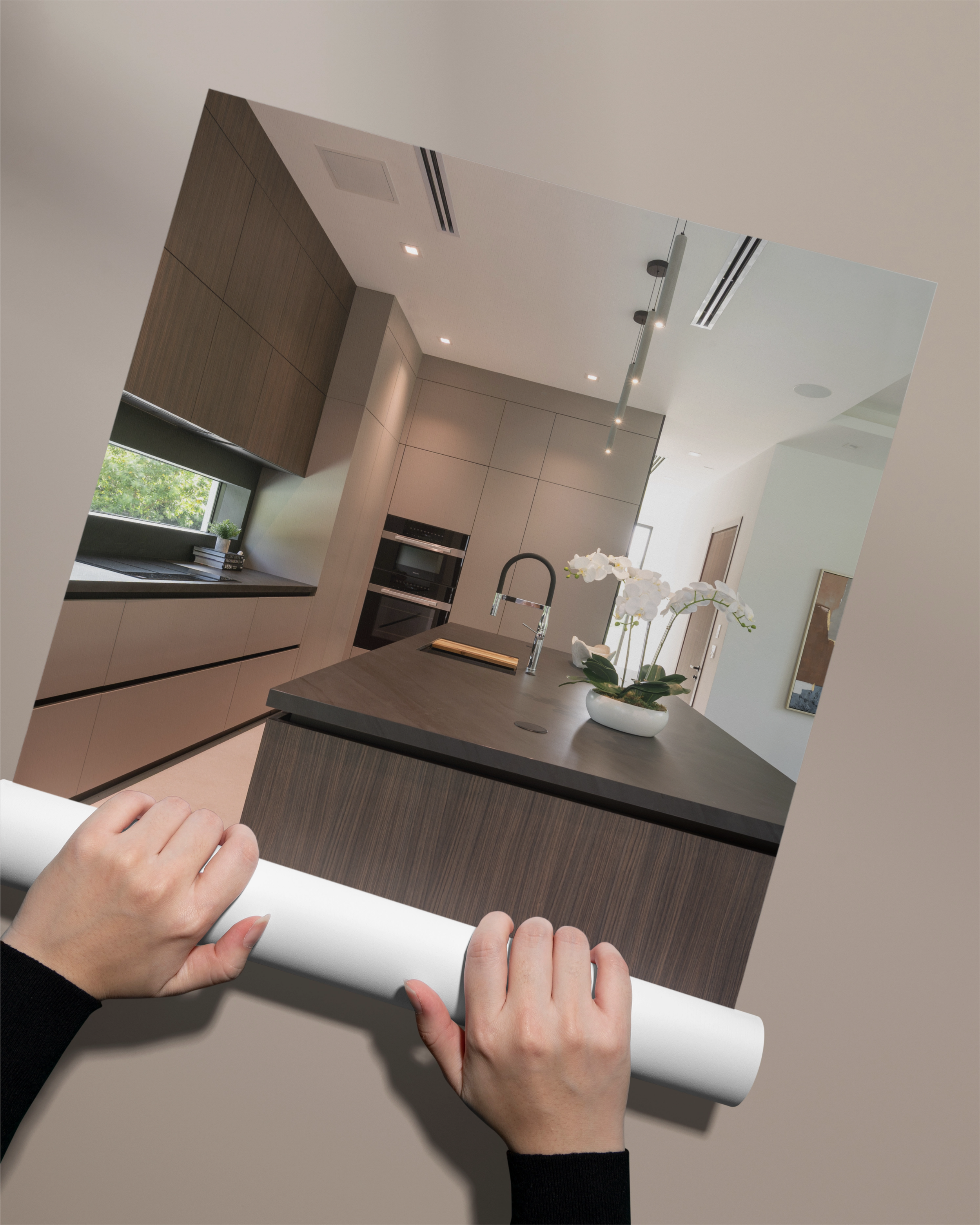 Person holding a large framed photo of a modern kitchen with wood cabinets, a black countertop, and a vase with white orchids on the island.