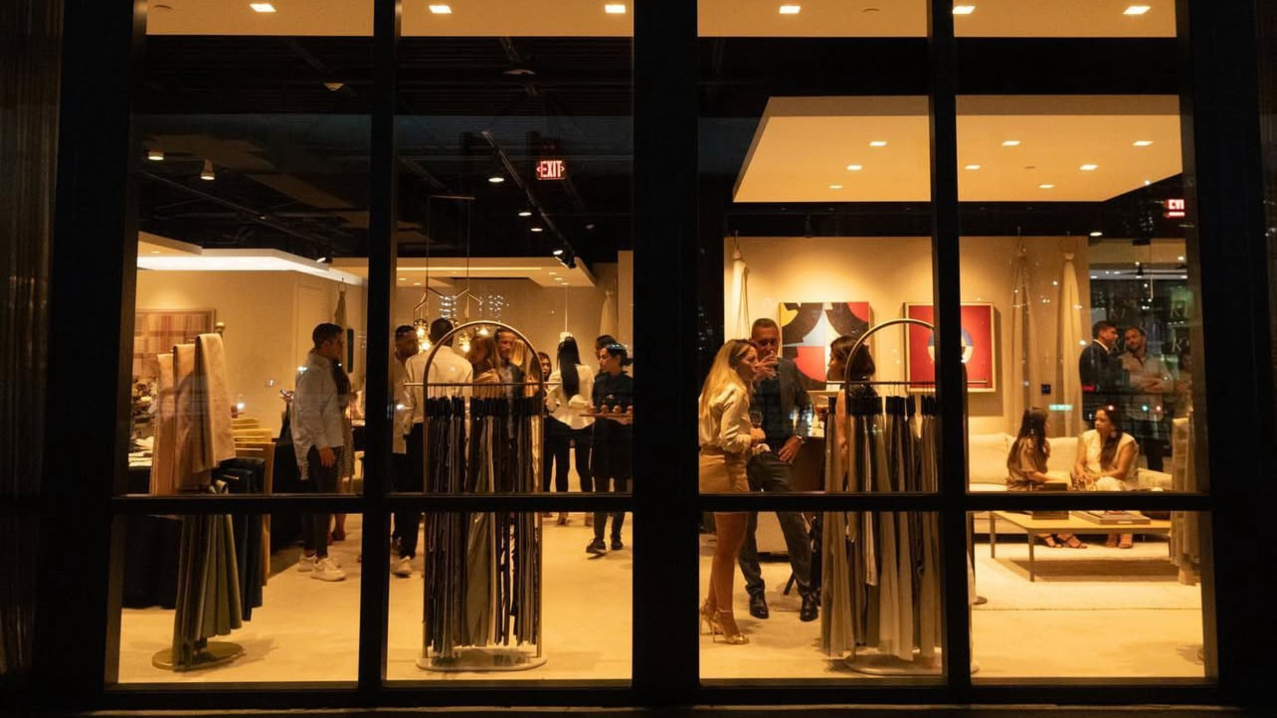People inside a modern, well-lit store viewing and discussing various clothing and home decor items, seen through a window at night.