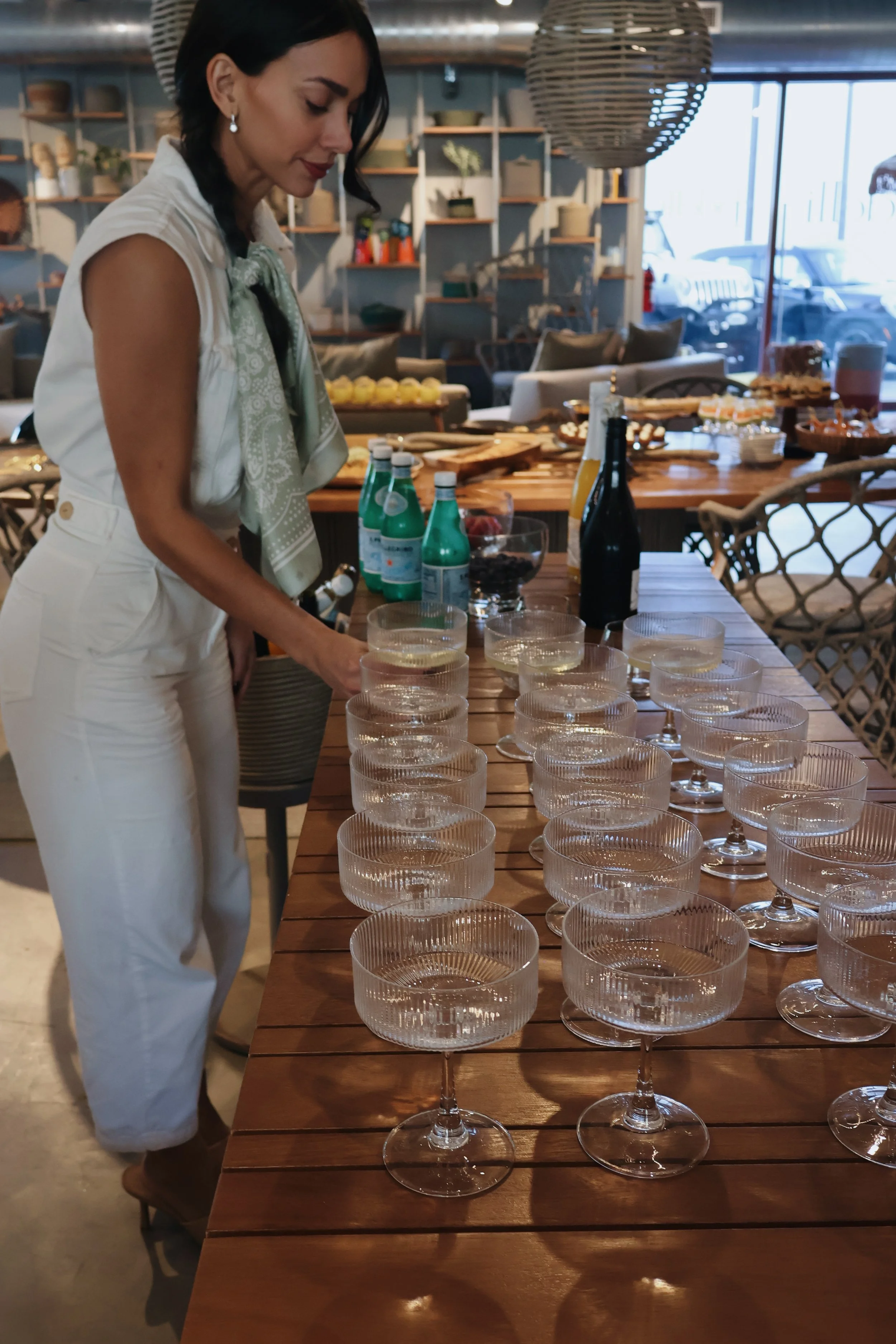 A woman in white pants and a sleeveless top, with a green scarf, arranging glasses of water on a wooden table in a cozy indoor space with shelves, chairs, and large windows.