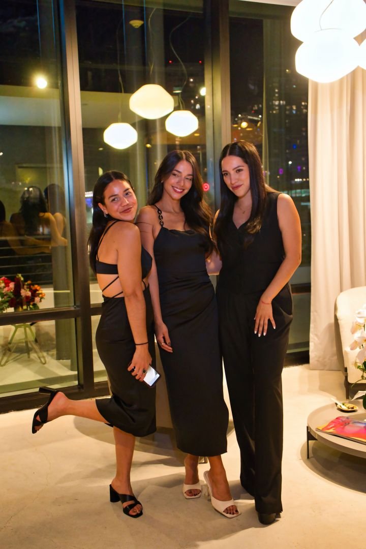 Three women in black dresses posing together indoors at night with large window behind them showing city lights, bright hanging lights above, and a white curtain to the right.
