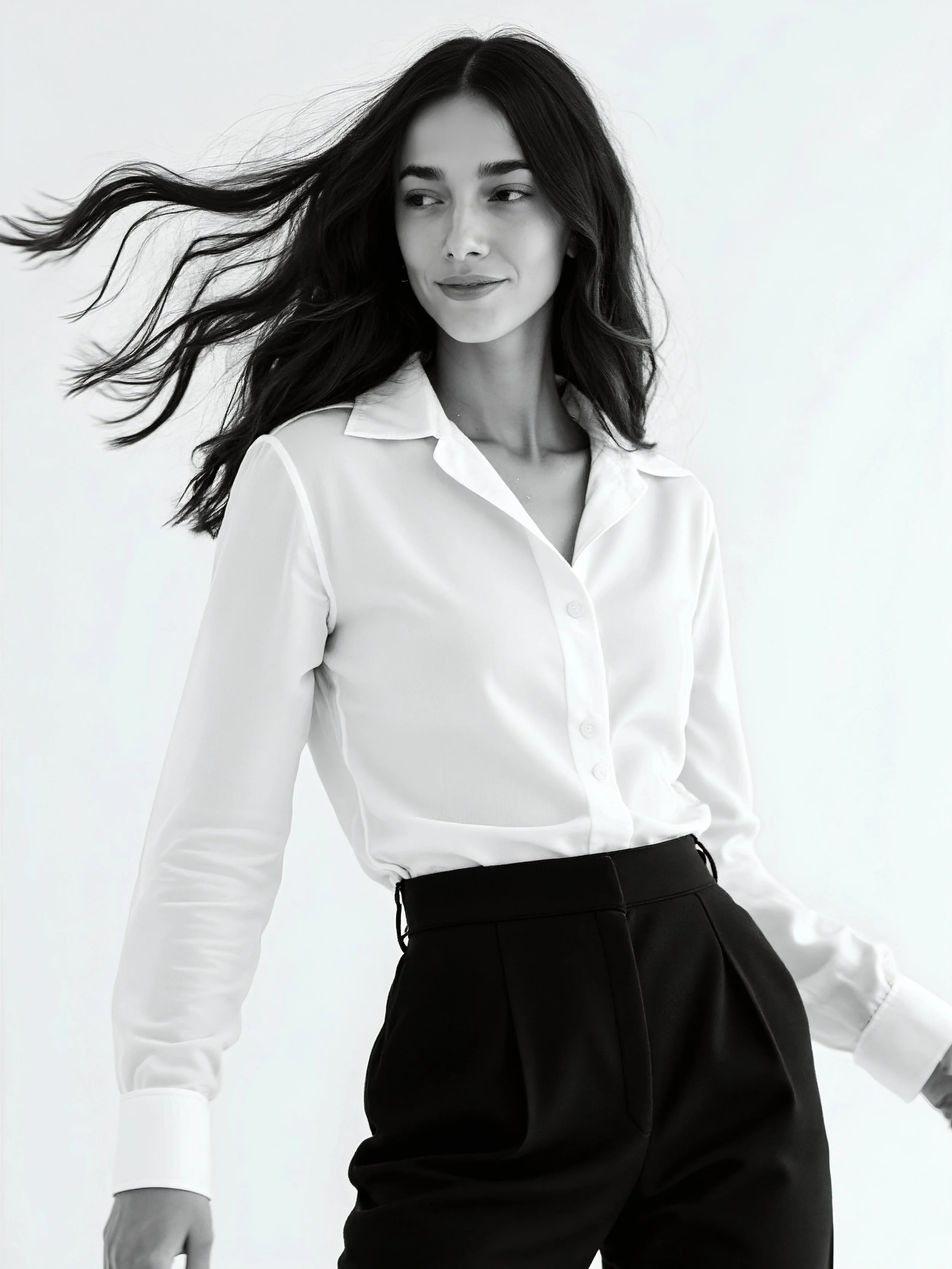 A woman with long dark hair wearing a white blouse and black high-waisted pants, standing against a plain background.