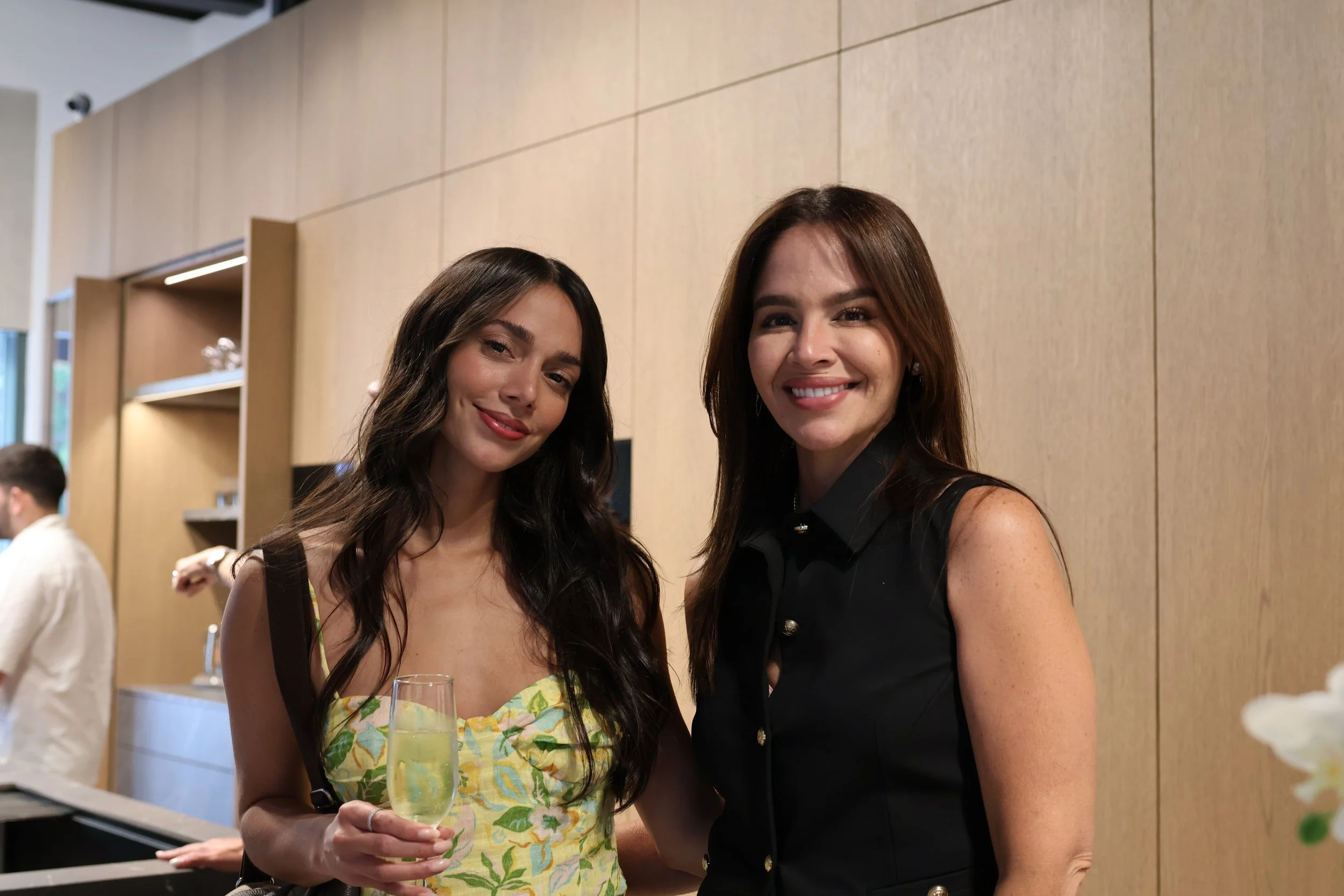 Two women smiling at camera at social gathering, one holding a glass of green drink, with a wooden-paneled wall in the background.