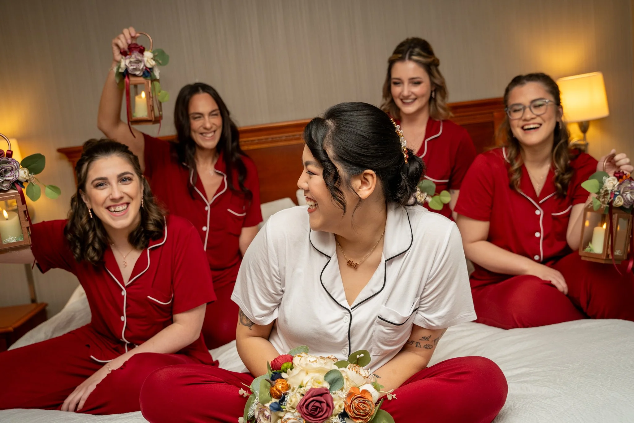 Bride and bridesmaids smiling together on a bed while holding lanterns during getting-ready moments at a Massachusetts wedding.