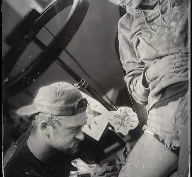 A man wearing a baseball cap sitting at a piano, with a woman standing nearby, showing a close-up of their lower bodies in a casual setting.