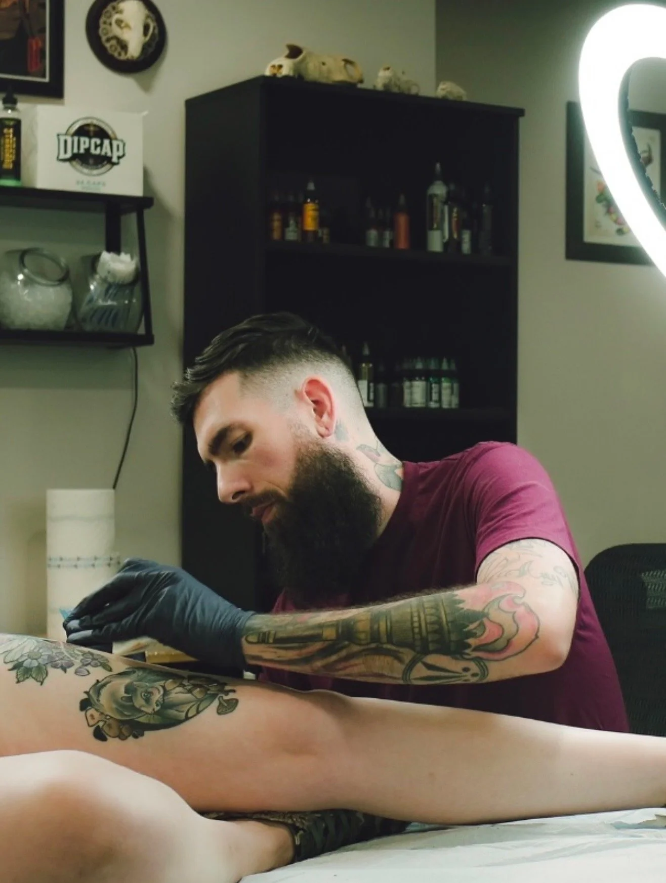 A man with tattoos and a beard getting a tattoo done on a person's leg in a tattoo studio.