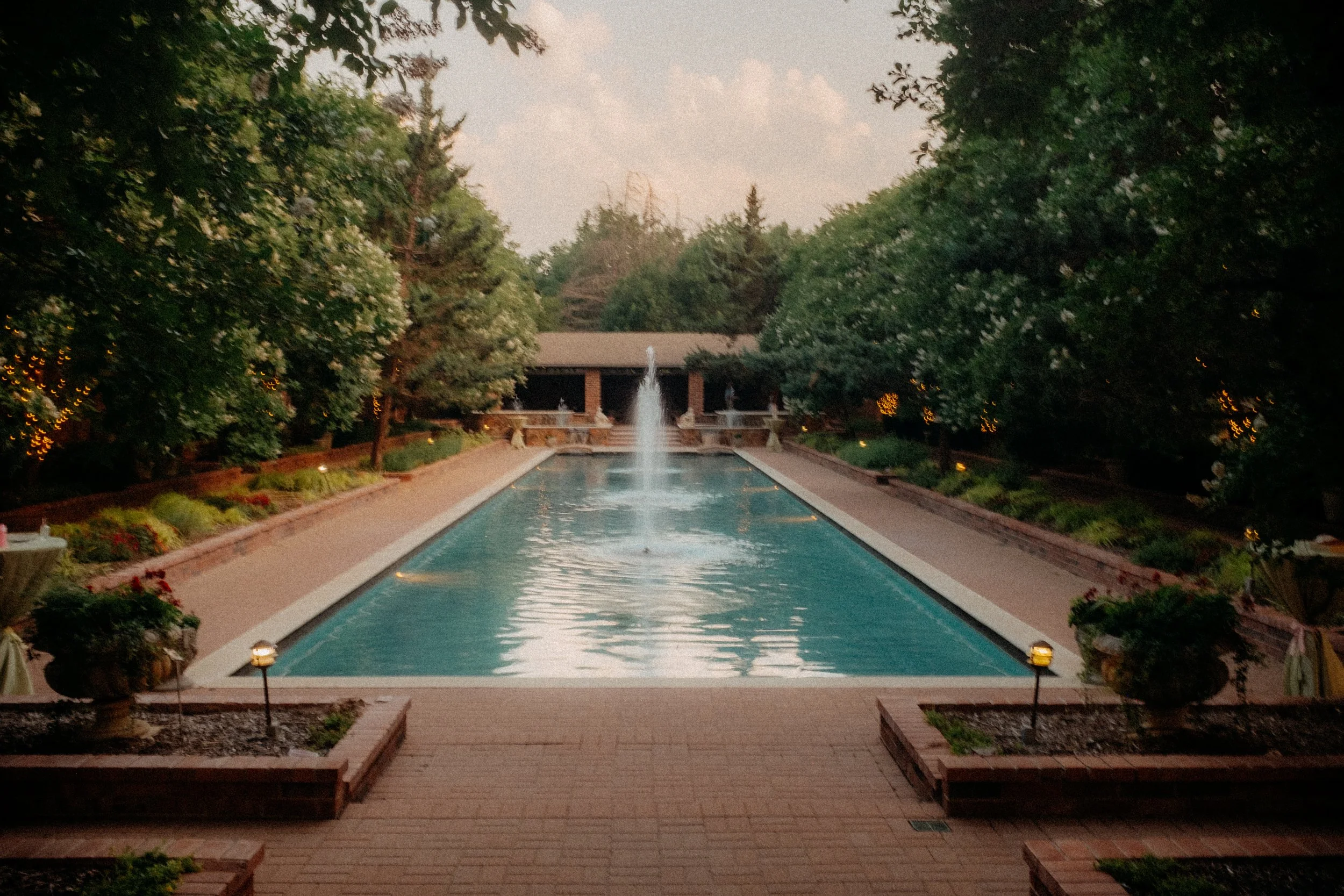 A tranquil outdoor swimming pool with a fountain at the center, surrounded by trees and landscaped gardens at dusk.
