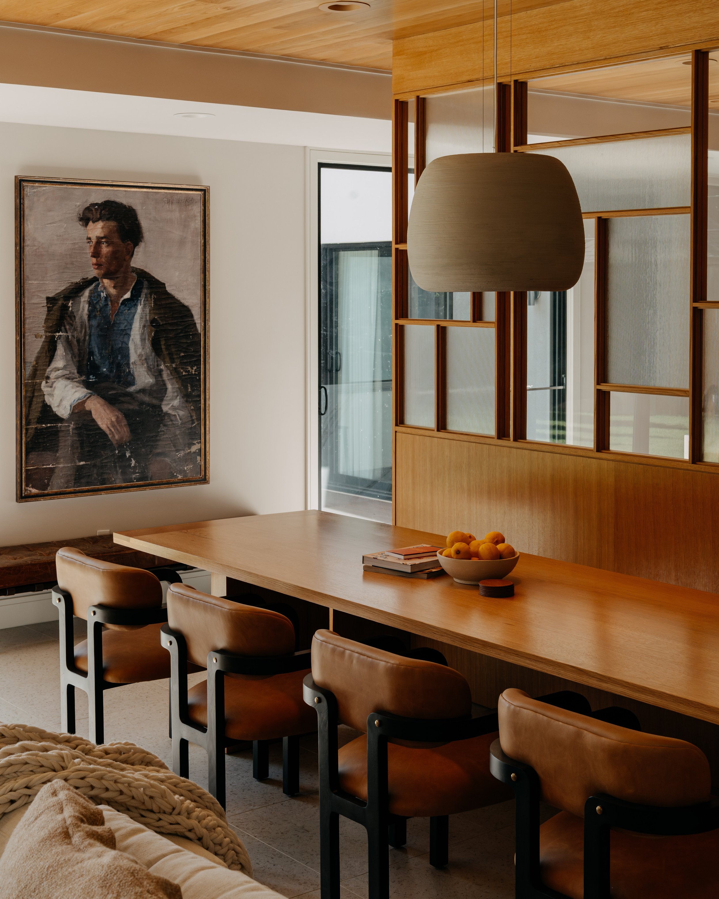 Modern dining room with a wooden table, six brown leather chairs, a bowl of peaches on the table, and a large portrait painting on the wall. Glass sliding door in the background, wooden ceiling, and partial view of a cream-colored sofa with chunky kn