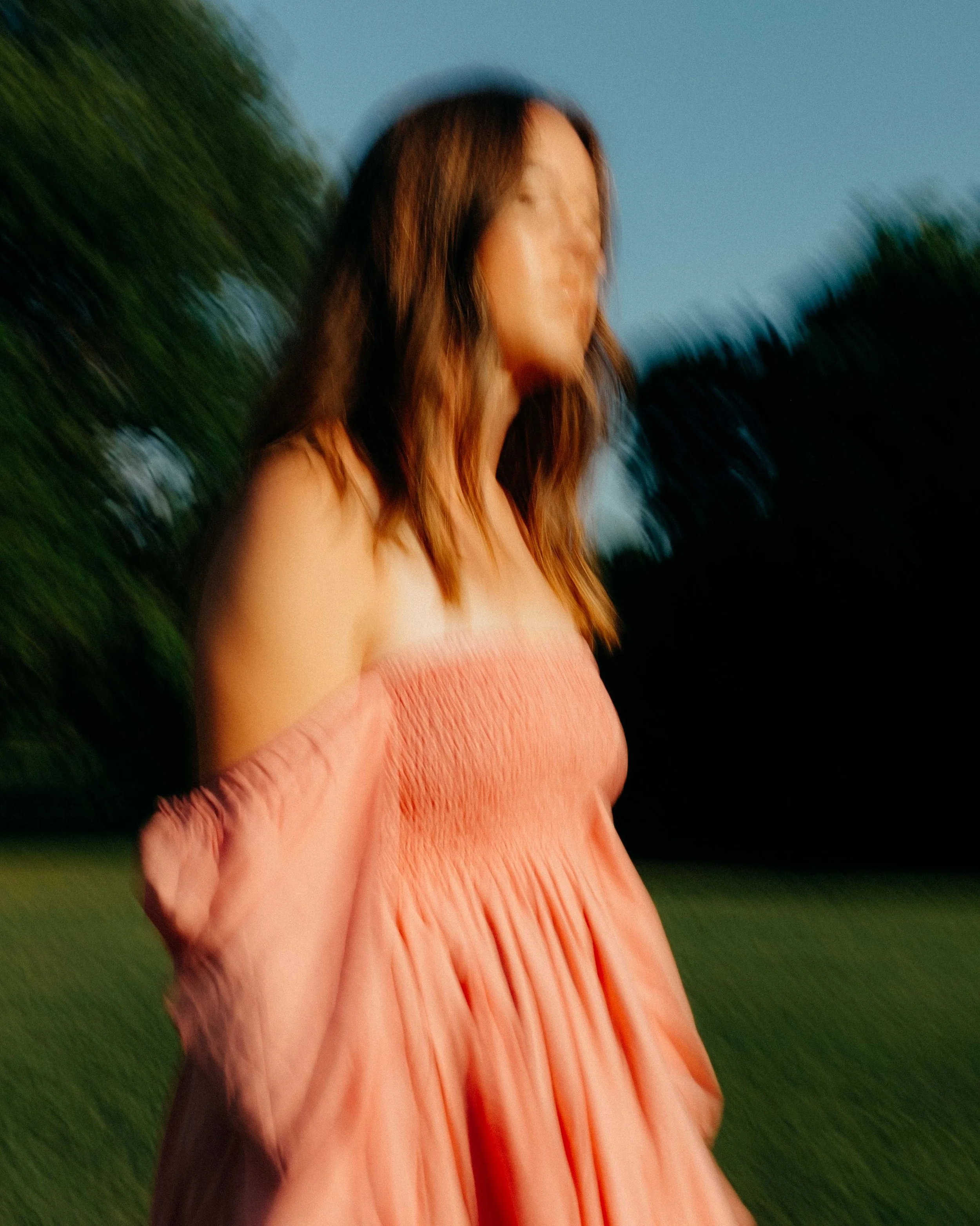 Blurry image of a woman in a peach-colored dress standing outdoors at dusk, with trees in the background.