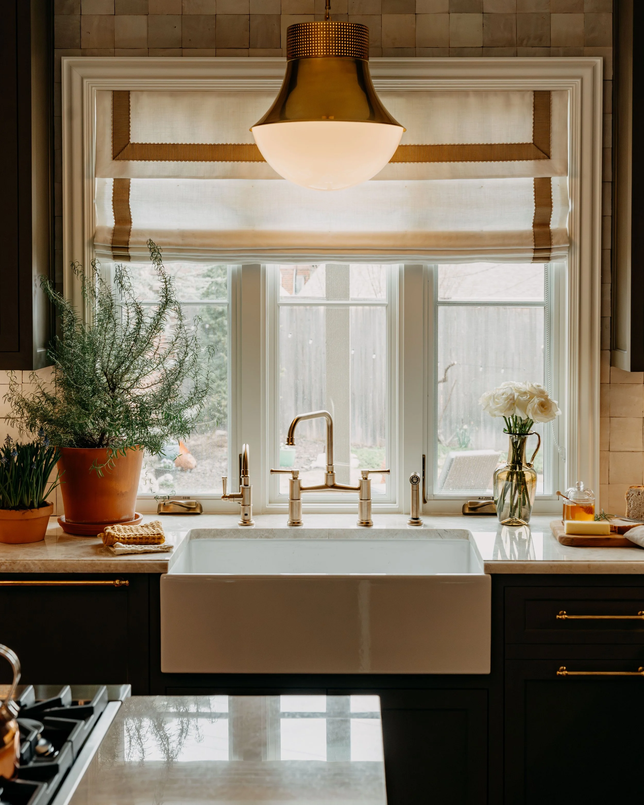 Kitchen sink area with a large window above, potted plants on the countertop, and a hanging light fixture.