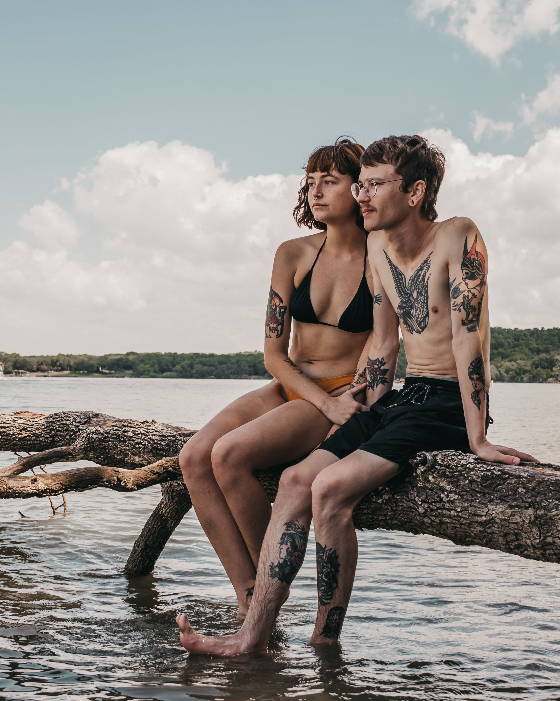 A young couple with tattoos sitting on a fallen tree branch in the water by a lake, relaxing on a cloudy day.