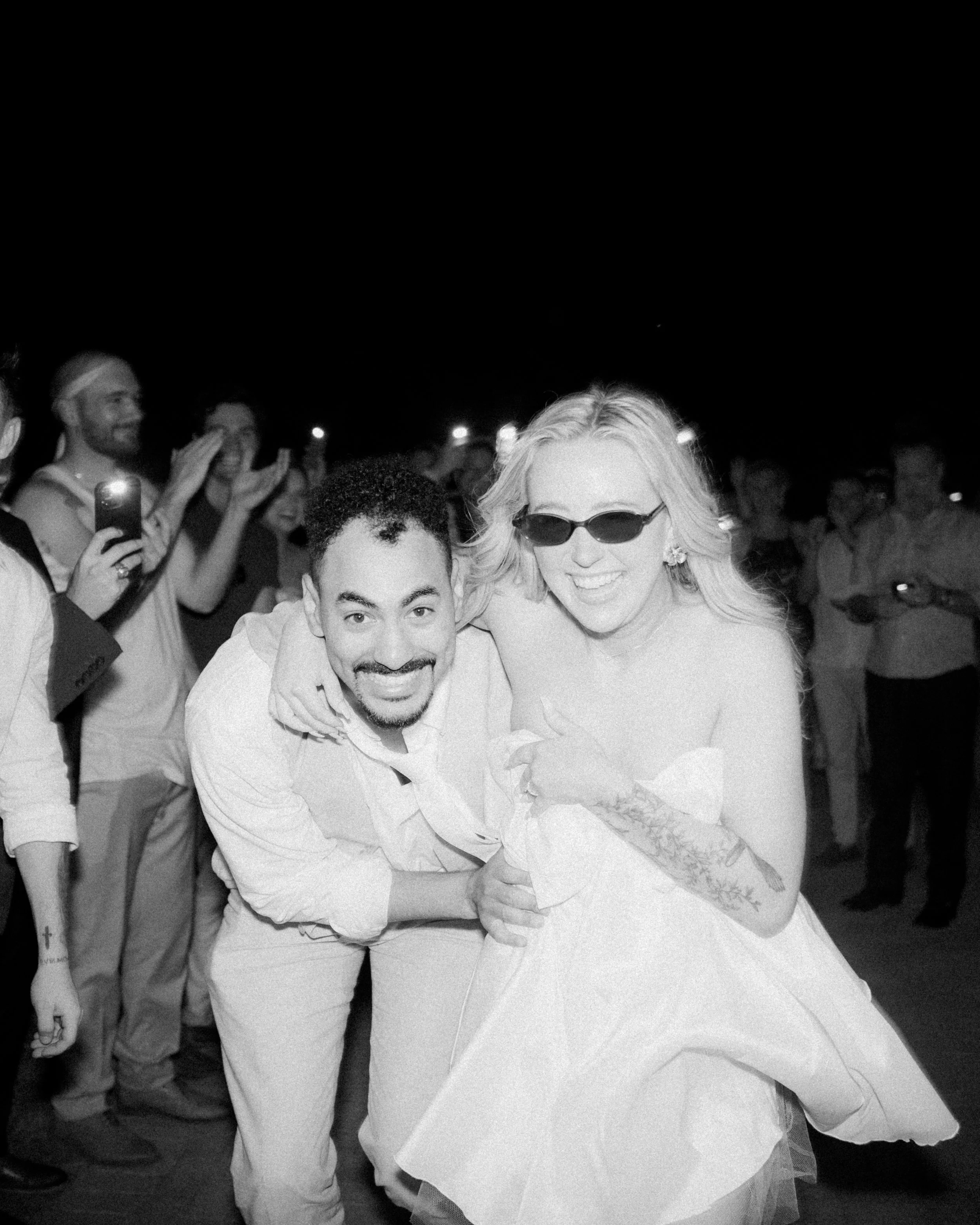 A black and white photo of a smiling couple at a celebration, with the woman wearing sunglasses and a strapless dress, surrounded by an appreciative crowd.