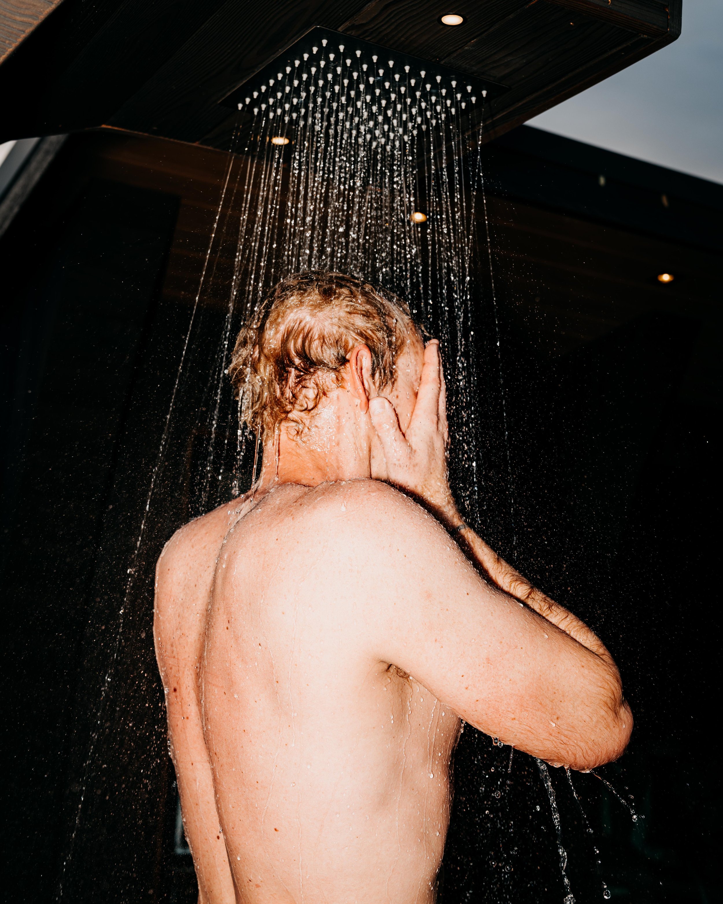 A man standing under an outdoor shower with water flowing down, covering his face with his hands.
