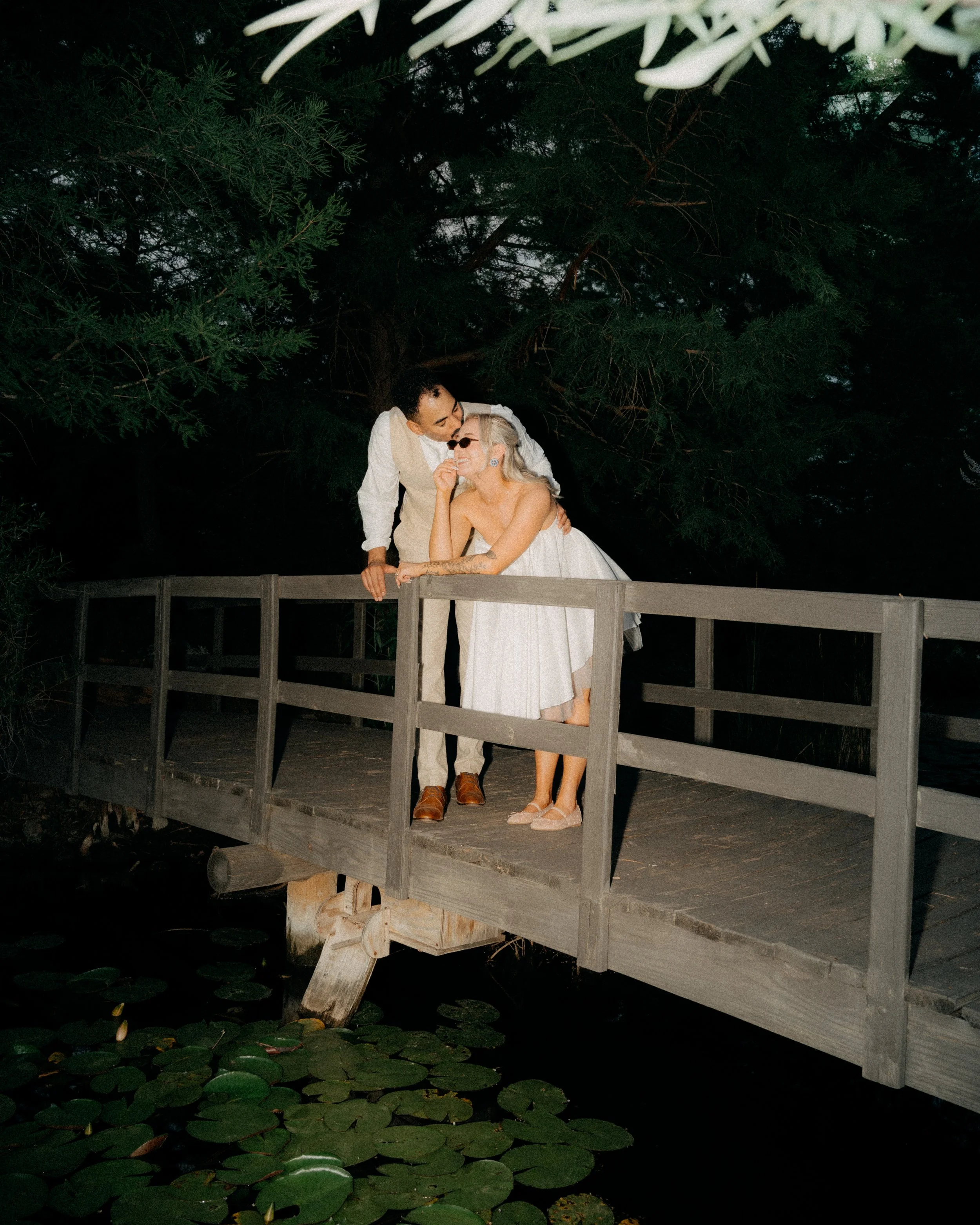 A couple dressed in wedding attire standing on a wooden dock over water with lily pads, sharing a moment together at night.
