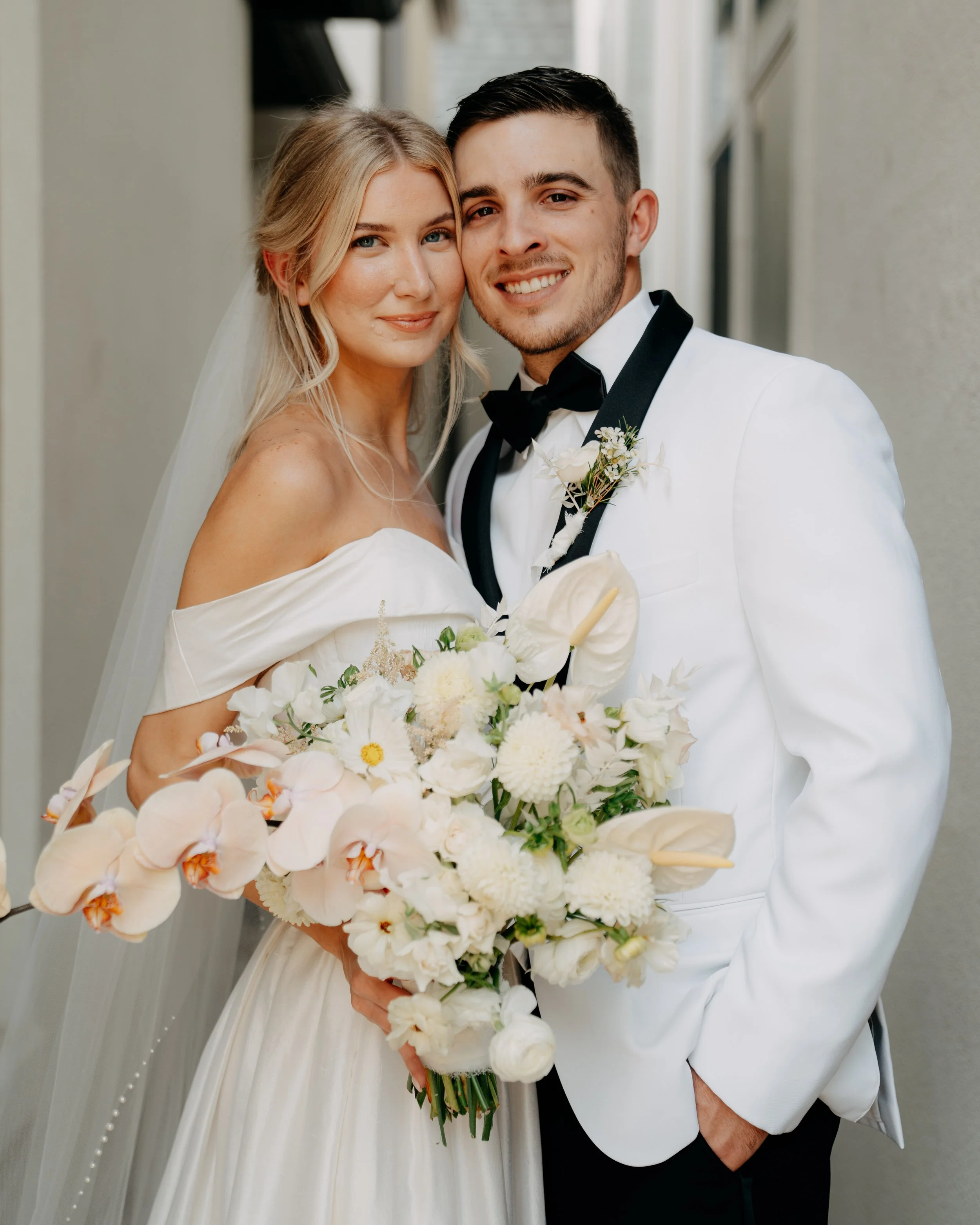 A bride and groom smiling and posing for a wedding photo, with the bride holding a bouquet of white and light pink flowers.
