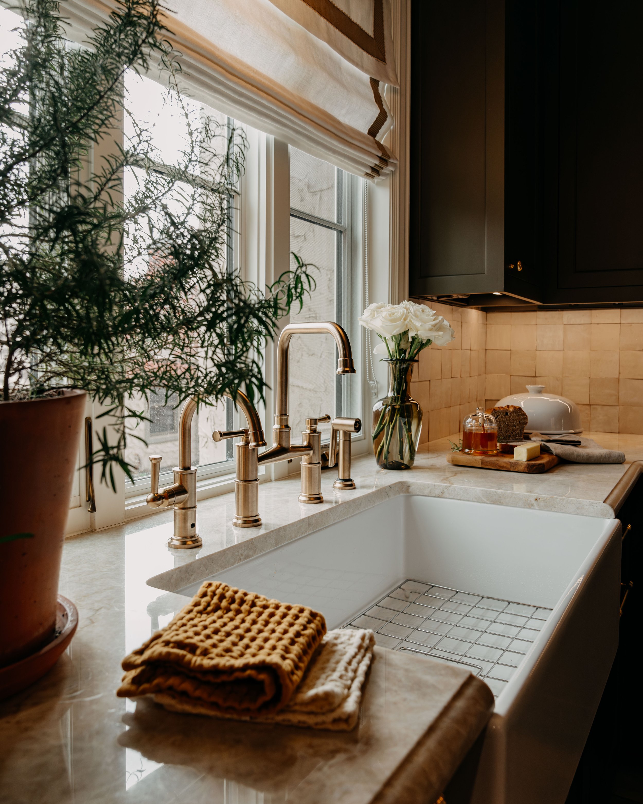 Kitchen countertop with a white farmhouse sink, brass faucet, beige and white towels, a potted plant, a vase with white roses, and various baked goods and butter on a cutting board near the window with natural light.