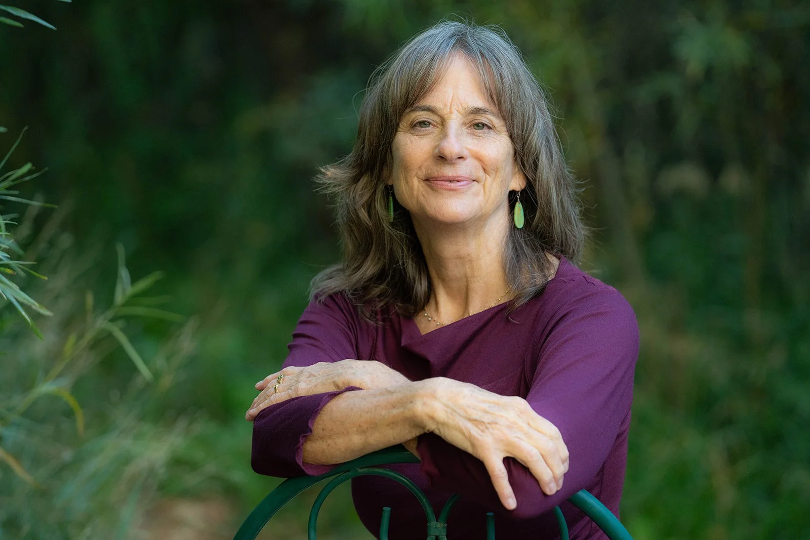 A middle-aged woman with gray hair wearing a purple top and green earrings, smiling outdoors with green foliage in the background, resting her arms on a green railing.