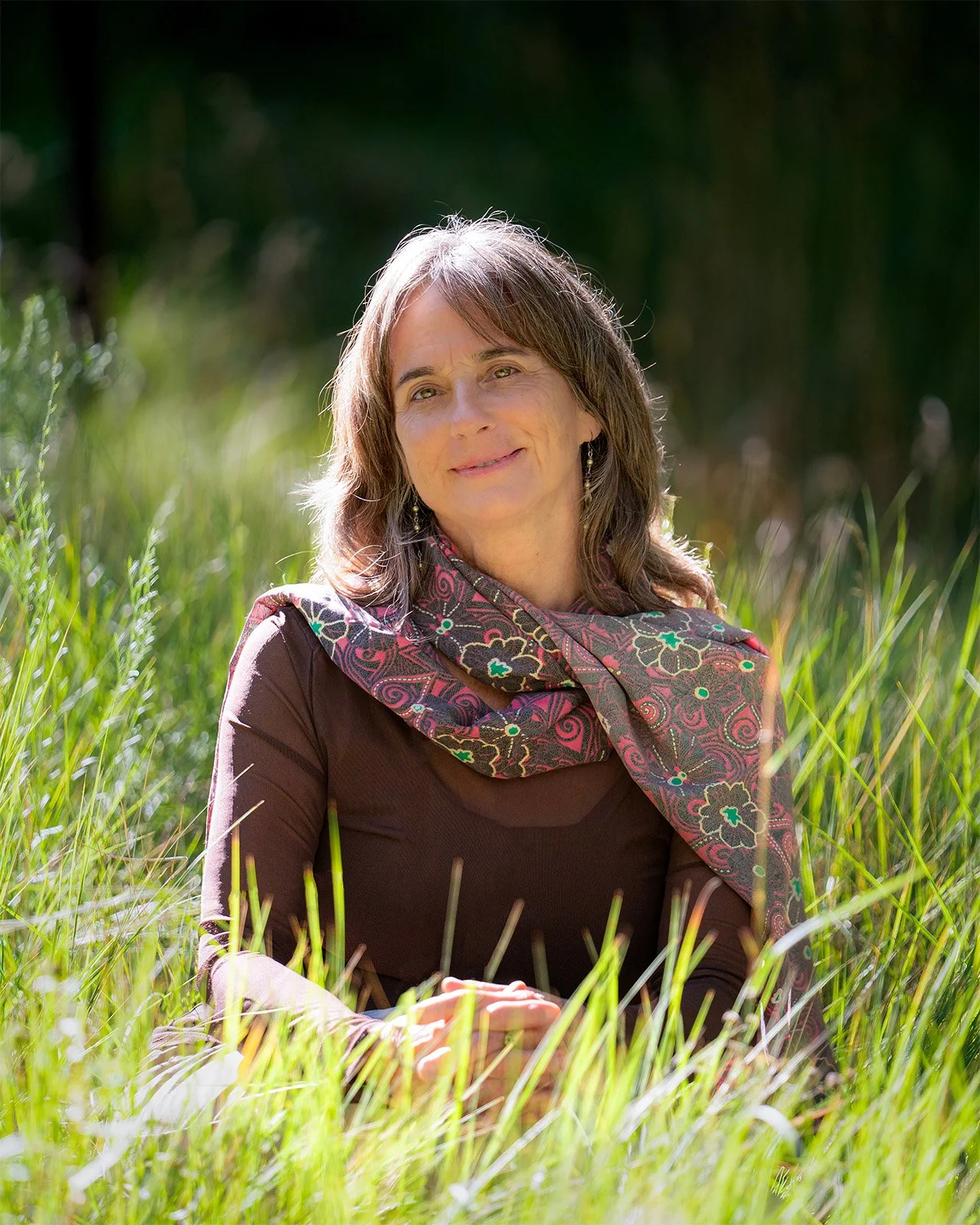 A woman with shoulder-length brown hair, wearing earrings and a colorful scarf, sitting outdoors in a grassy area with sunlight shining on her, smiling softly.