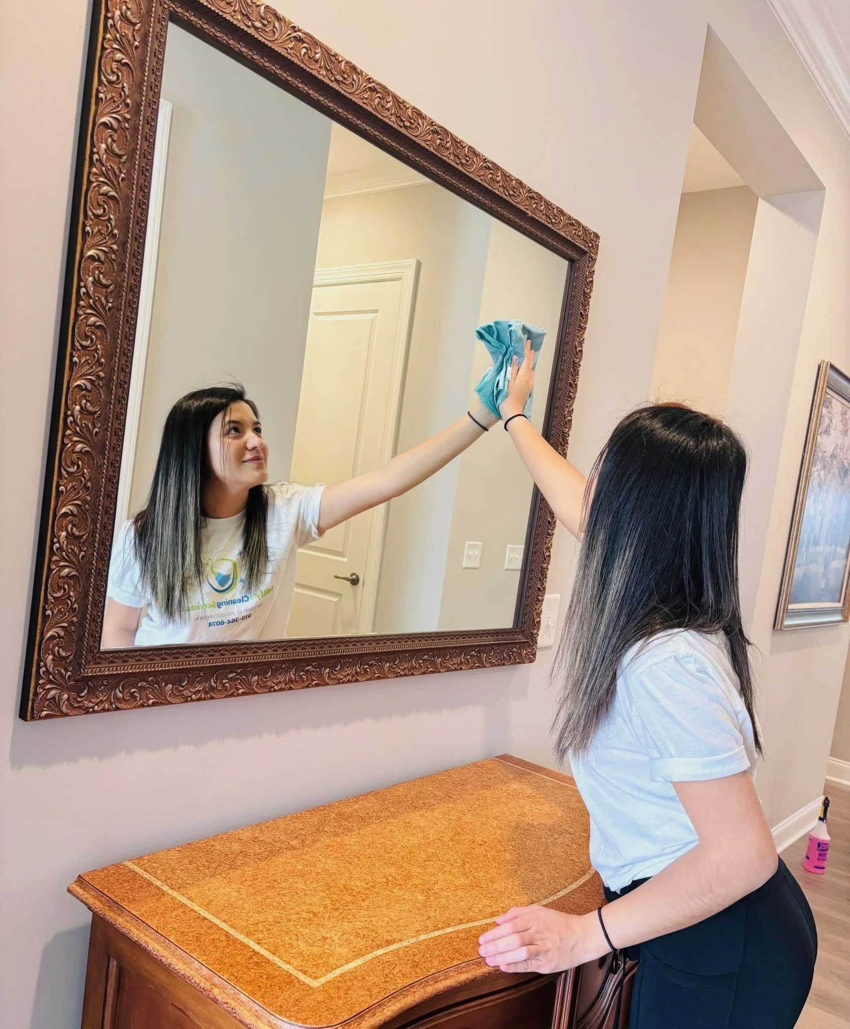 A young woman is cleaning a large mirror with a blue cloth while looking at her reflection in a room with light-colored walls and framed artwork.