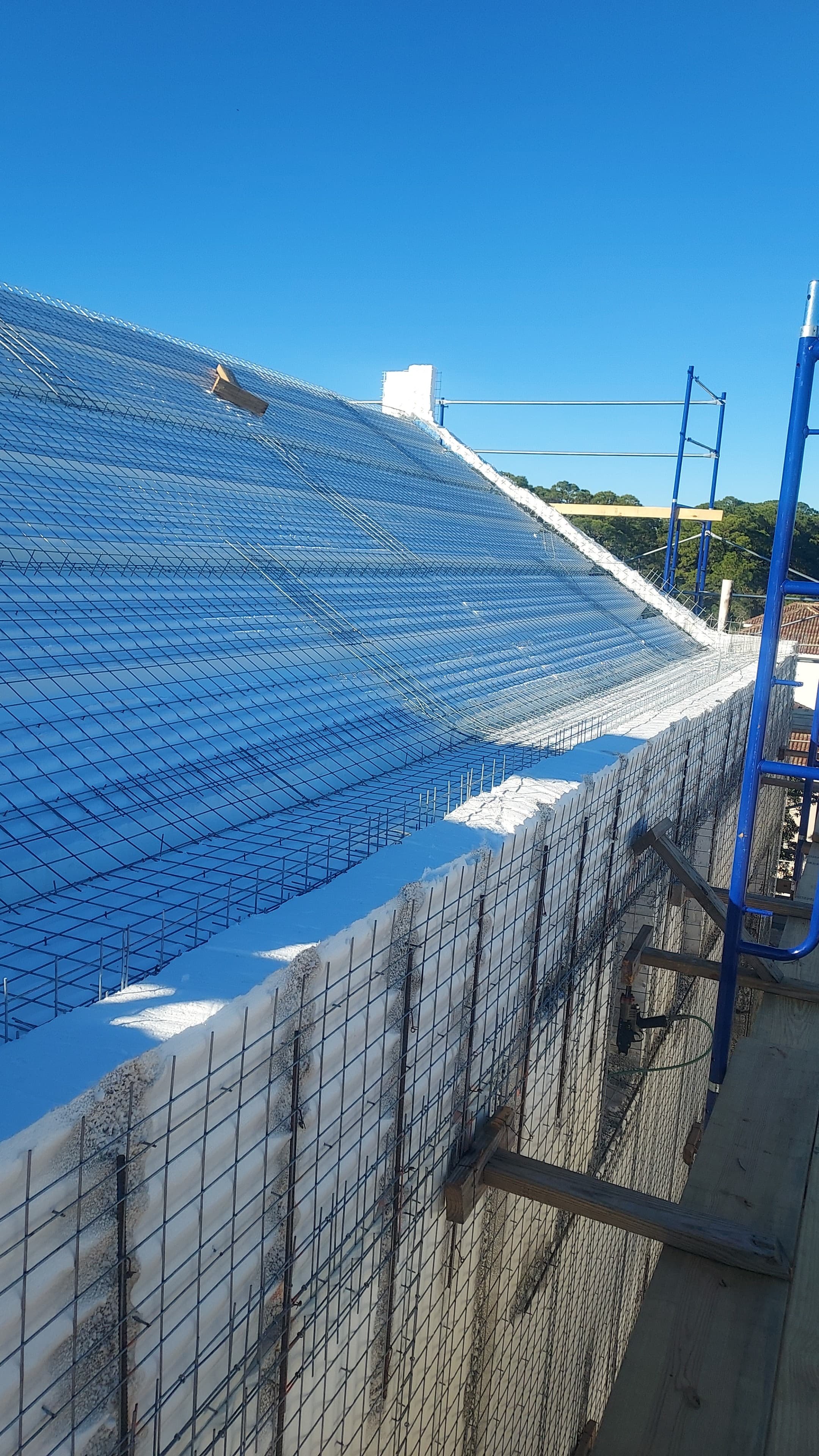 Construction site with a sloped roof covered in metal wire mesh and foam insulation, scaffolding is visible on the side, under a clear blue sky.