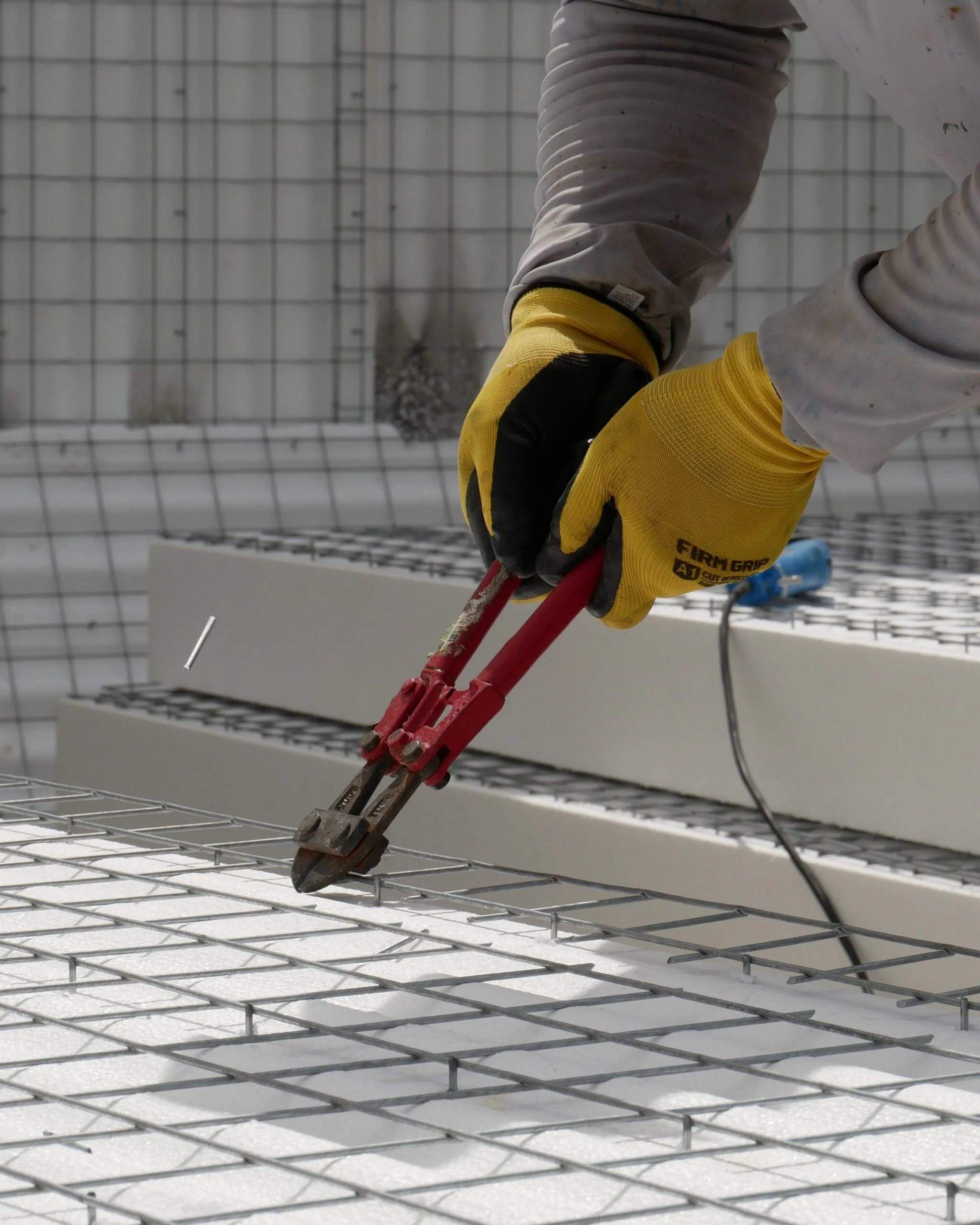 Person wearing yellow gloves and white long-sleeve shirt using red pliers to cut or work on metal wire grid in construction setting.
