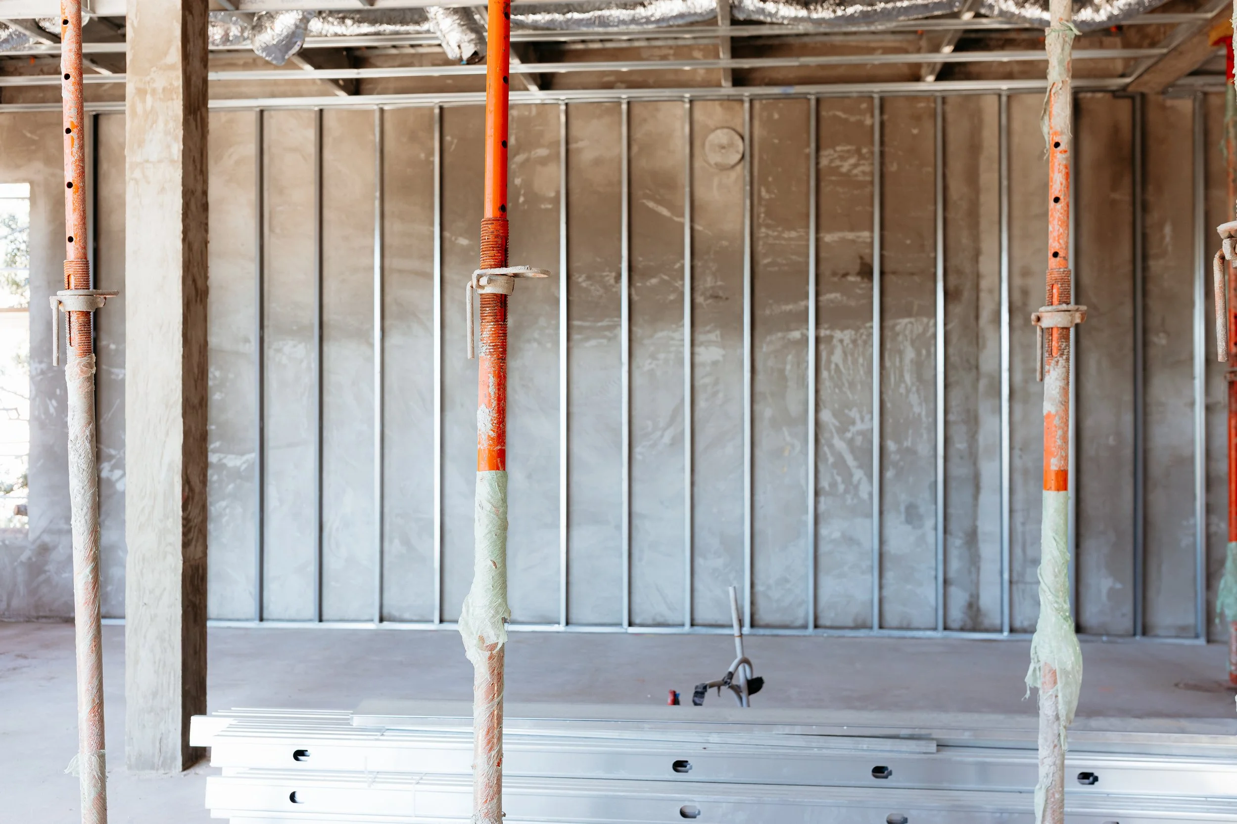 Interior view of a building under construction with metal framing, orange scaffolding poles, and a stack of metal studs on the floor.