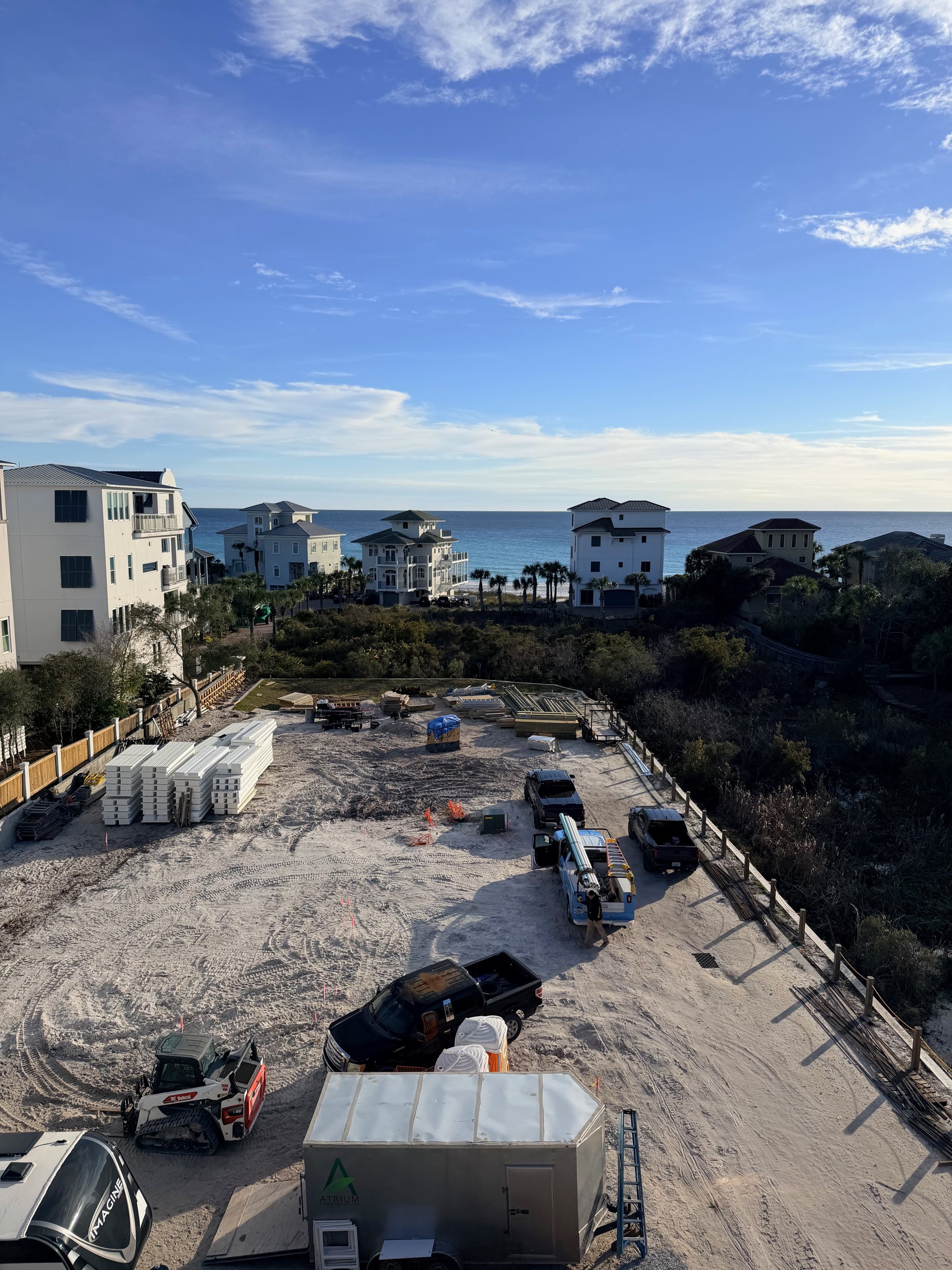 View of a beachside construction site with various vehicles and building materials, overlooking the ocean and adjacent coastal houses under a blue sky with scattered clouds.