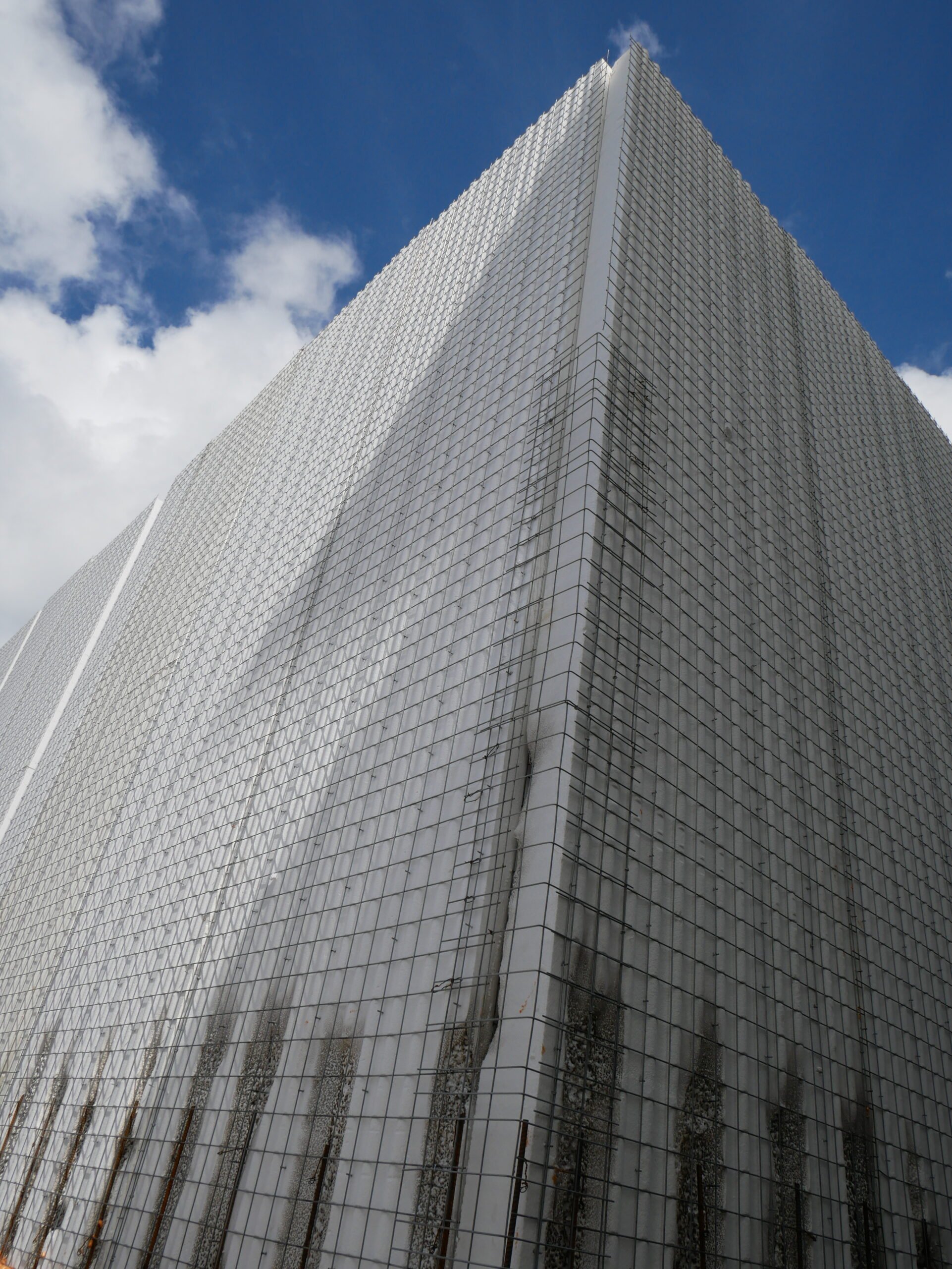A tall skyscraper with a glass exterior, reflecting the blue sky and clouds, showing signs of damage on the building's surface.