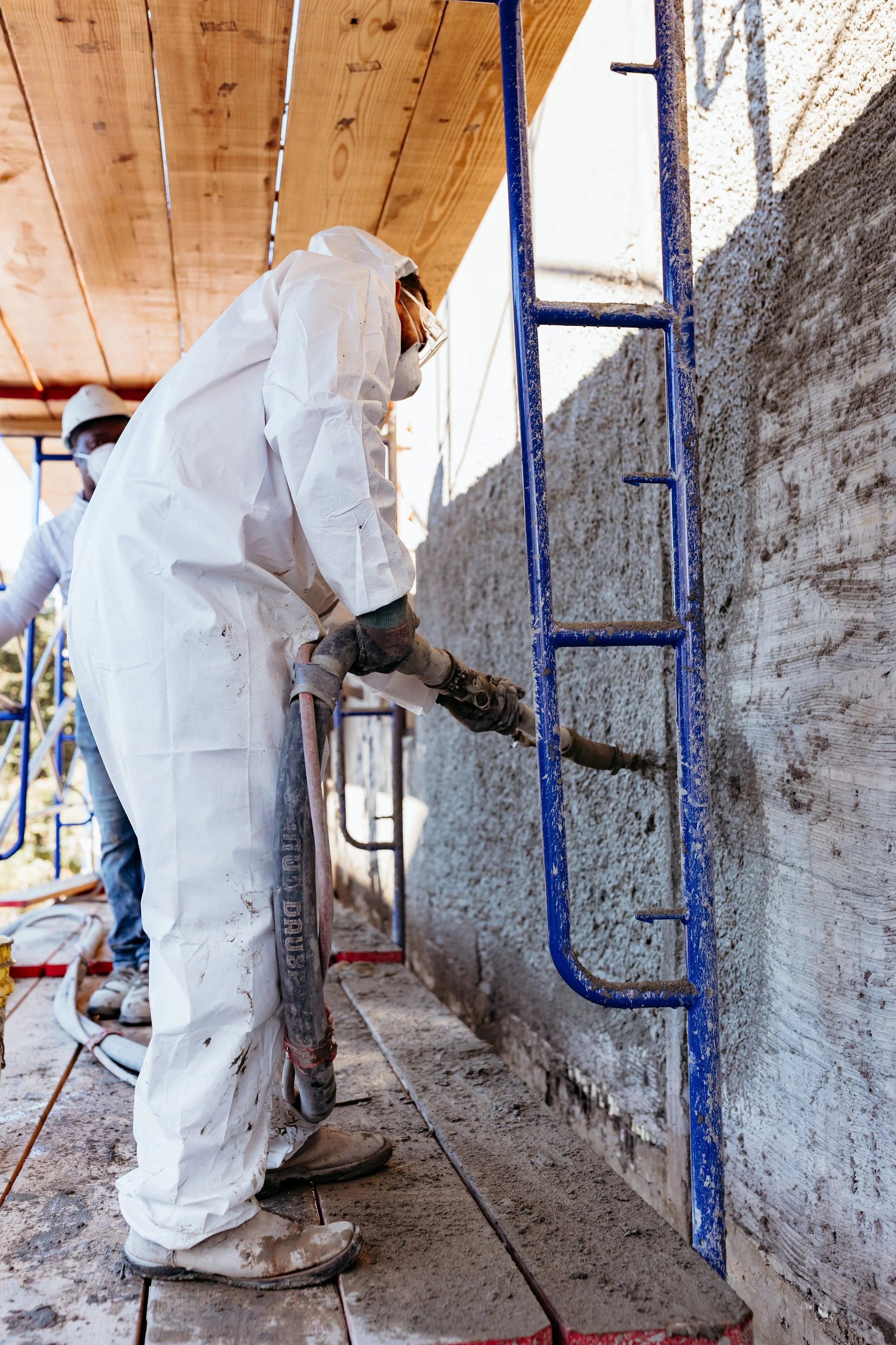 Worker in protective gear using a sprayer on a concrete wall, with scaffolding nearby.