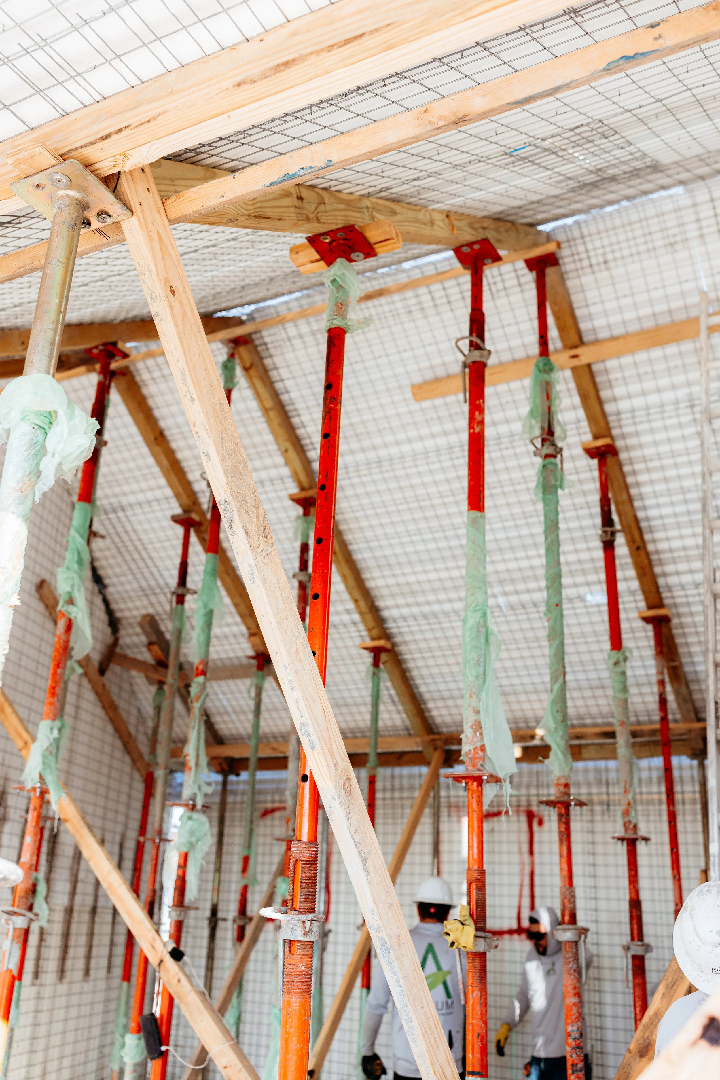 Construction site with orange and red scaffolding and wooden supports, two workers in safety helmets and gloves working inside.
