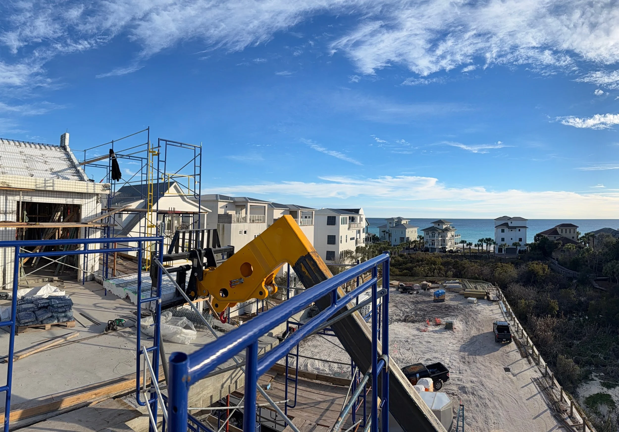 A construction site on a partly built residential building with a yellow crane arm, blue scaffolding, and construction materials, overlooking a neighborhood of beach houses by the ocean.