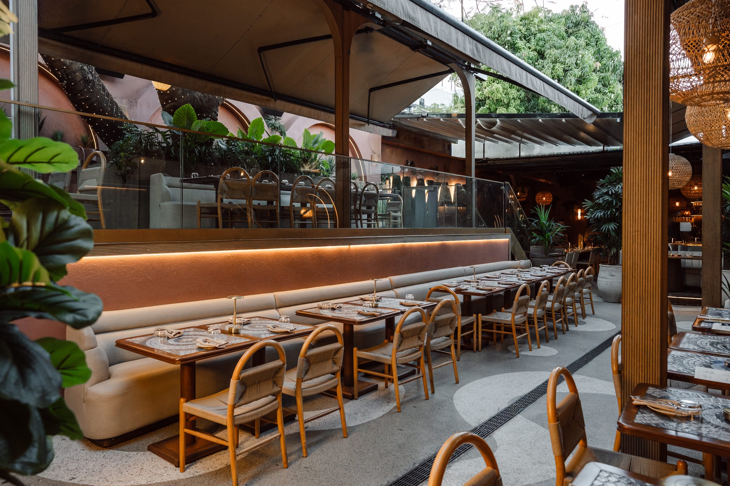 Empty indoor restaurant with wooden chairs, marble-topped tables, and lush green plants, featuring high ceilings with natural light and warm lighting fixtures.