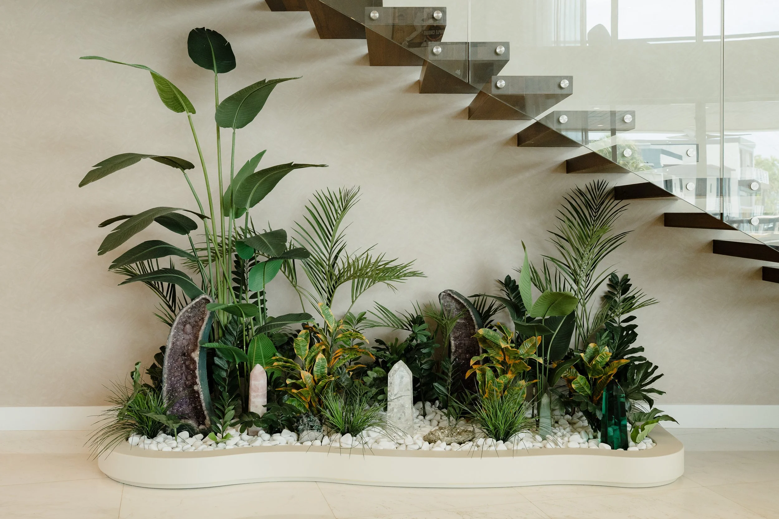 Indoor modern planting display with various green plants, crystals, rocks, and white stones in a white container, with a staircase and large window in the background