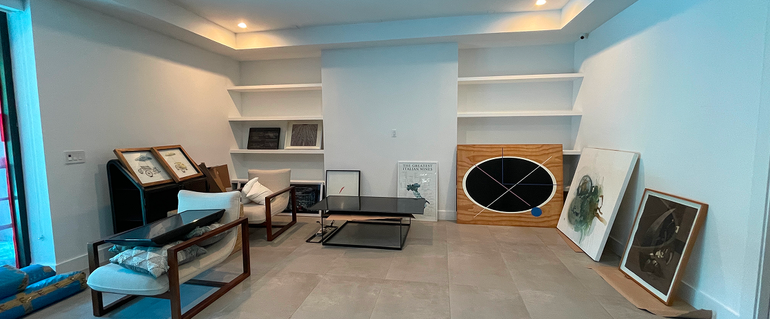 Living room with white walls and built-in shelves, featuring framed pictures, seating, and several large paintings leaning against the wall.
