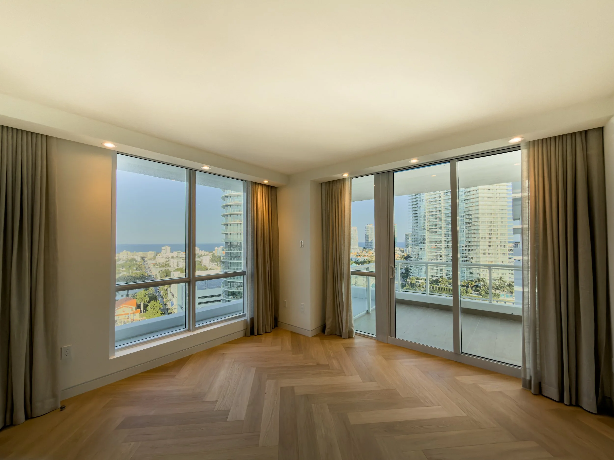 Living room with large windows showing city buildings, beige curtains, and wooden flooring.