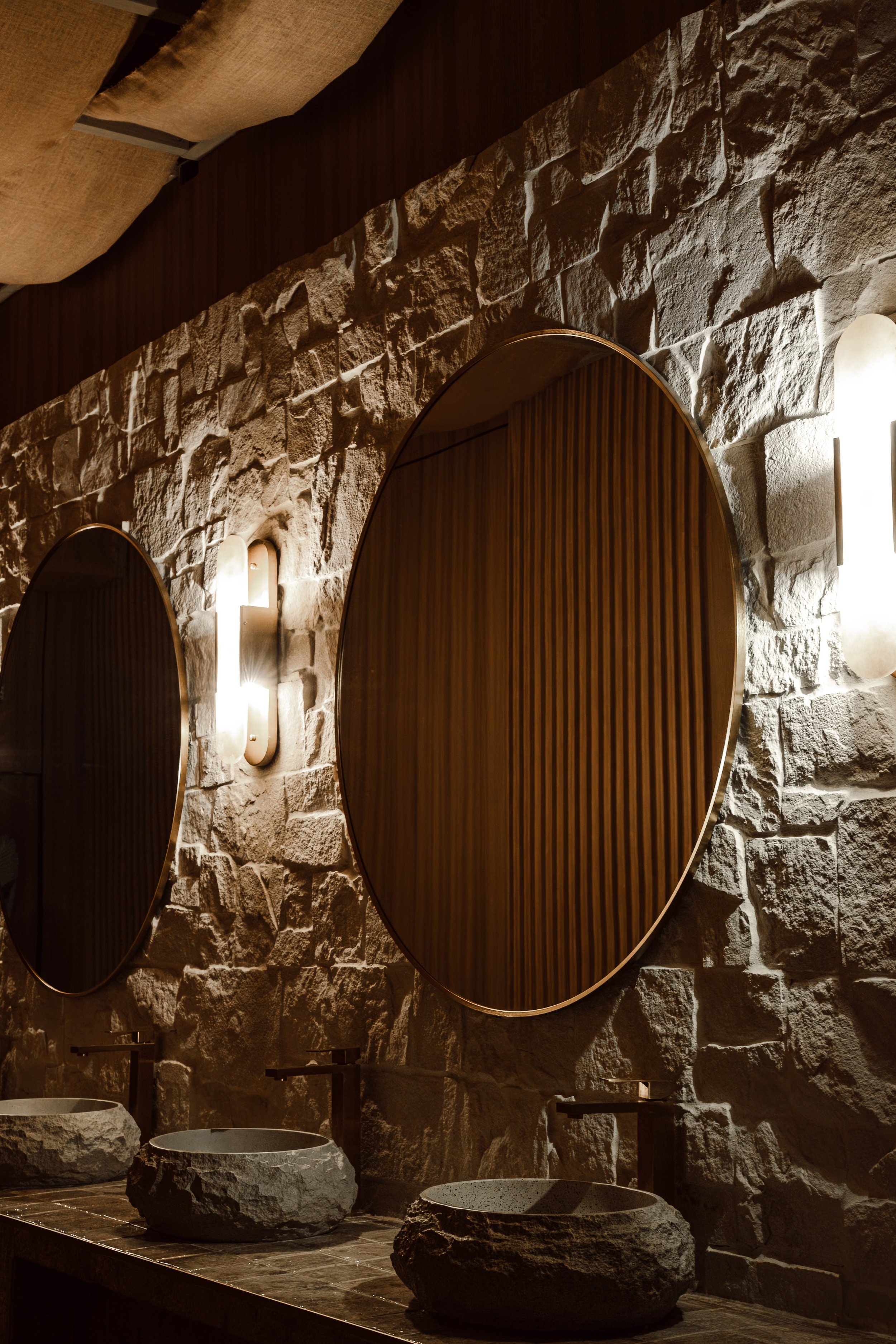 Stone wall with oval wooden mirrors and wall sconces, rustic bathroom with stone sinks.