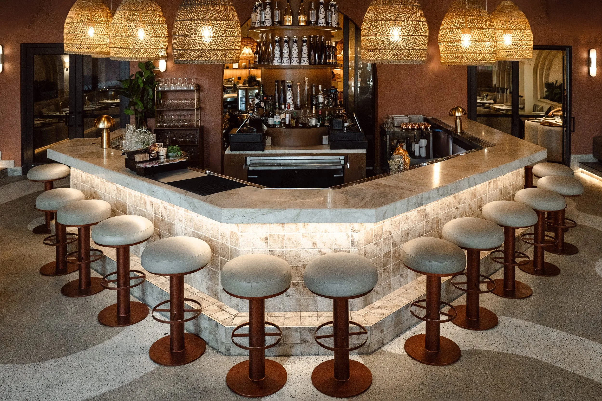 A circular bar with a marble top and cushioned stools around it, situated in a warmly lit restaurant or bar with wicker pendant lights hanging overhead.