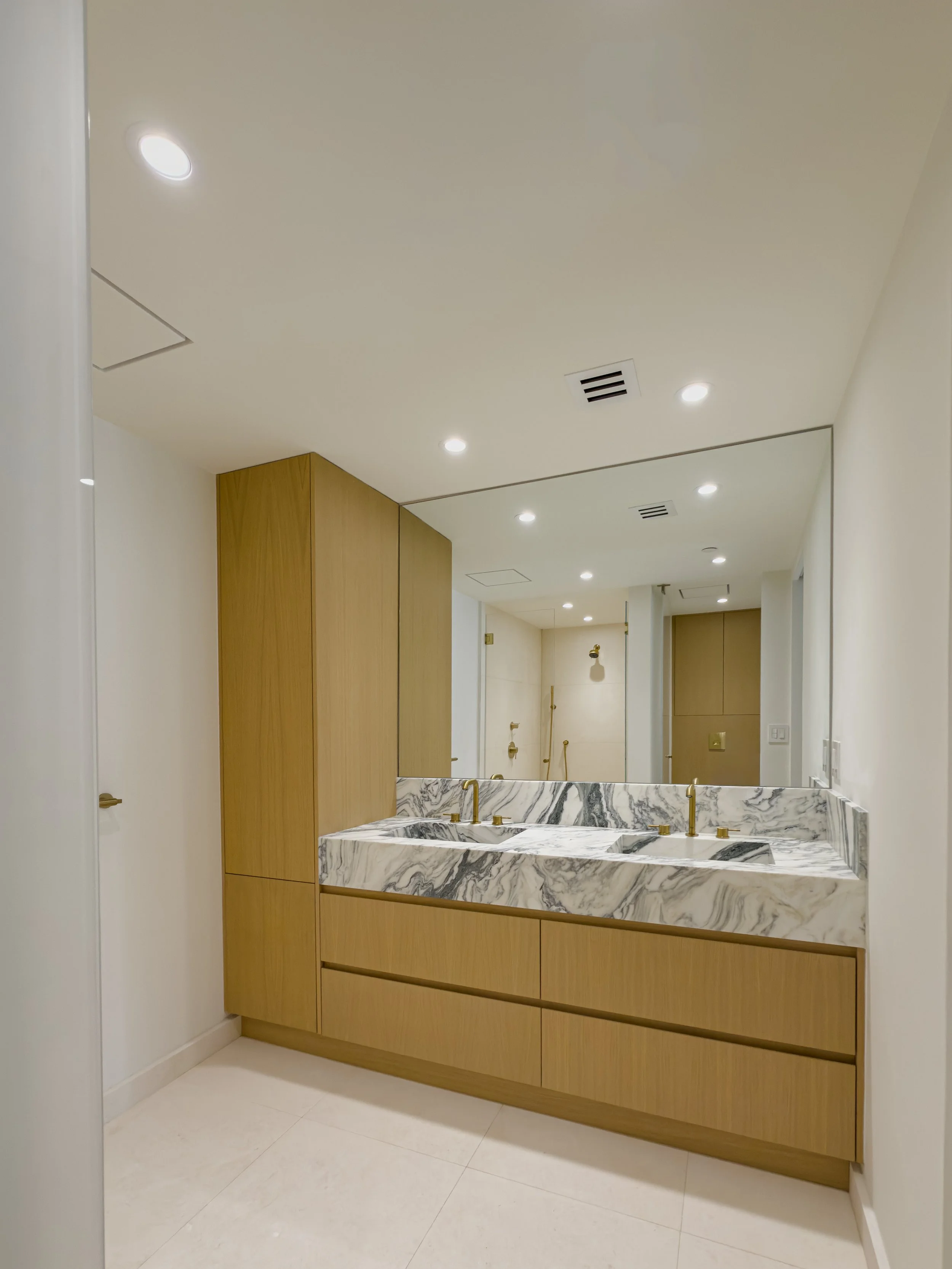 A marble double sink with gold faucets attached to a wooden vanity in a modern bathroom, with a large mirror and recessed ceiling lights.