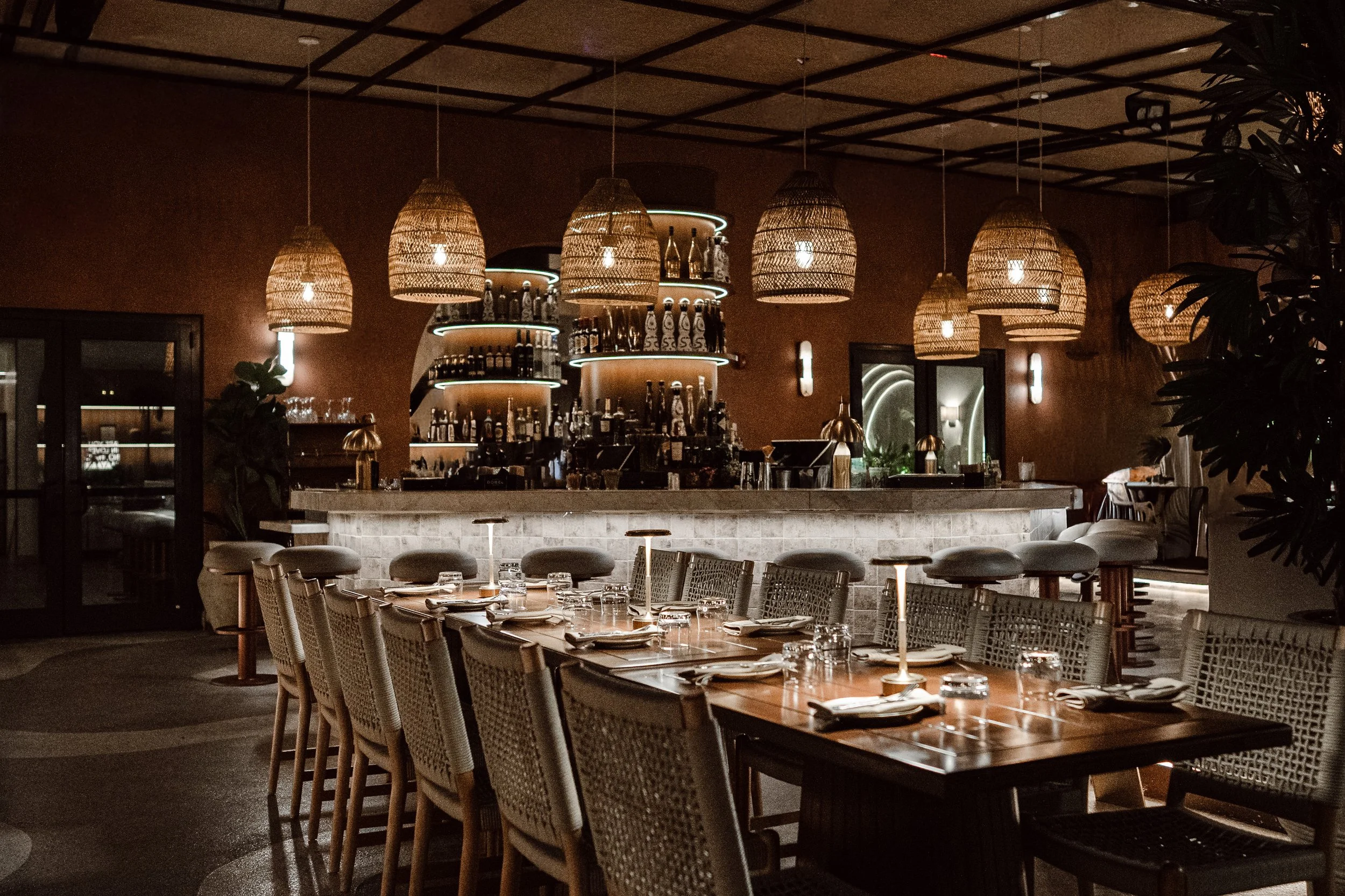 A dimly lit restaurant interior featuring hanging woven pendant lights, a bar with bottles, and a dining table set with glasses and plates.