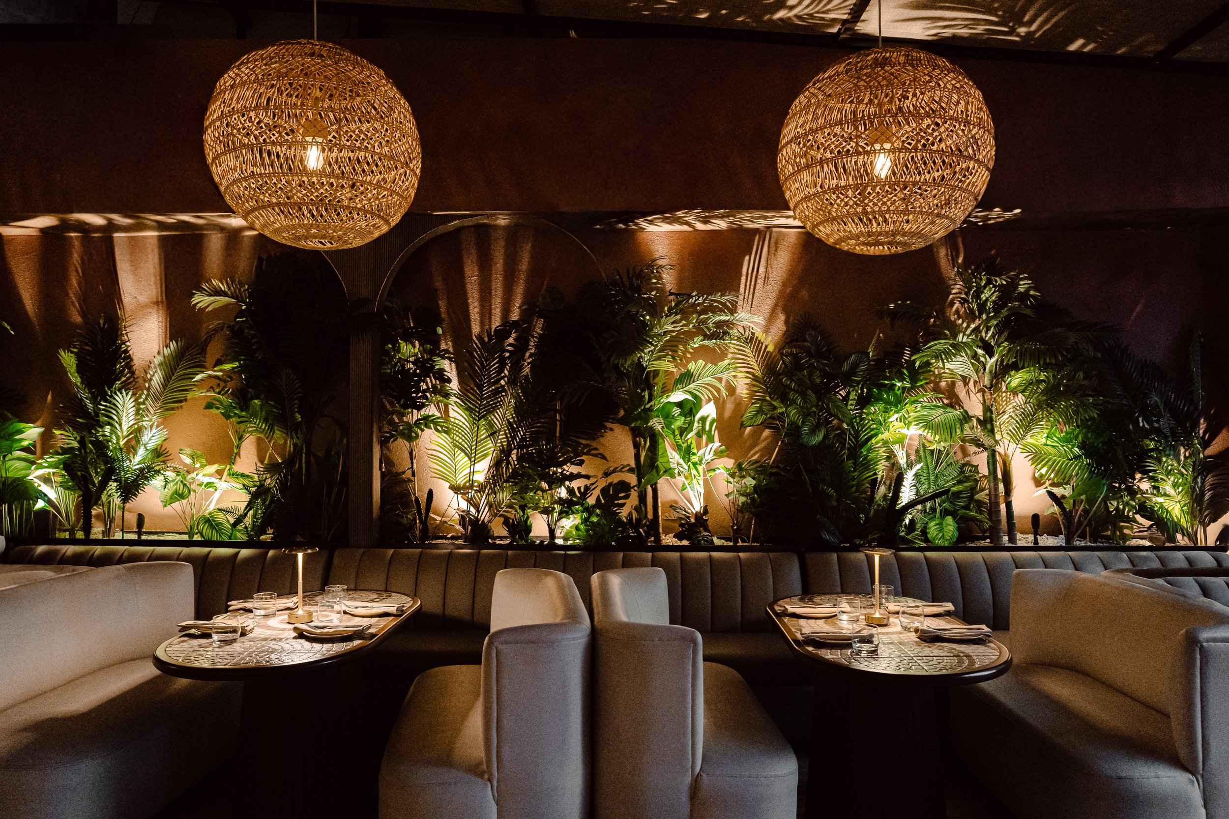 A dimly lit restaurant interior featuring two large woven pendant lights hanging from the ceiling, a lush-green plant arrangement against the wall, a long curved seating area with white upholstered chairs, and tables set with plates, glasses, and sil